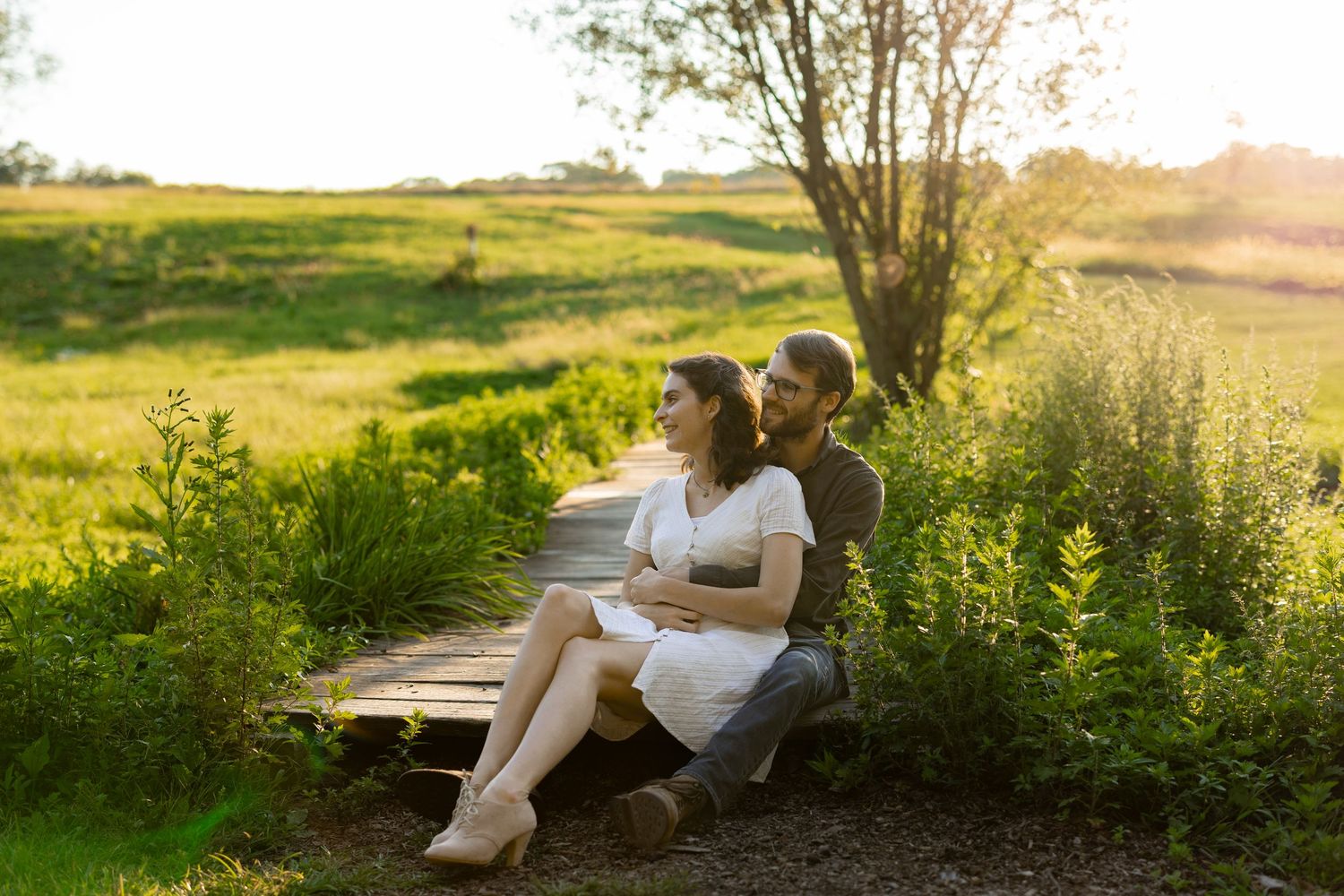 Couple in white clothing sits together on grassy hillside during golden sunset hour.