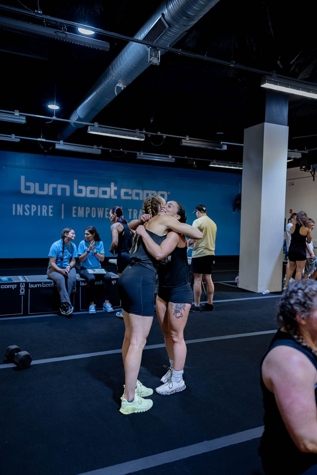 Two people share an emotional embrace during a fitness class at Burn Boot Camp with blue wall branding visible.