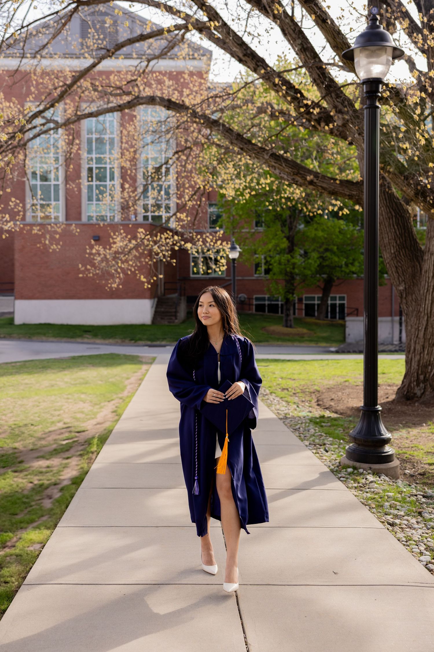 Graduate in navy blue commencement gown walks along campus pathway near brick building on sunny spring day.