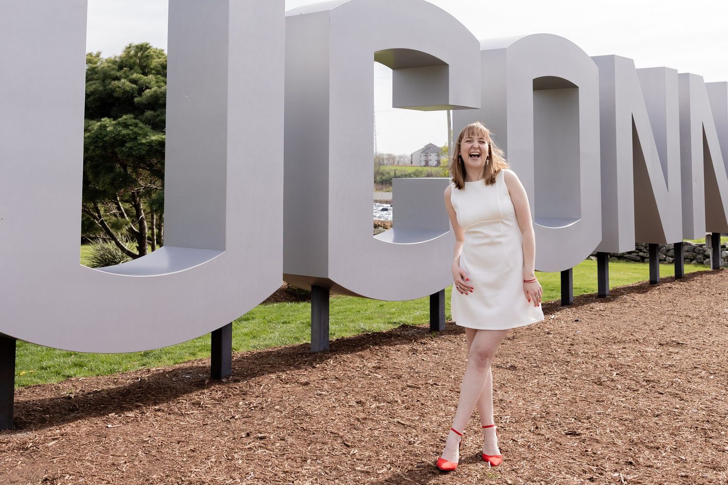 Person in a white dress poses in front of large white letters at an outdoor location during daytime.