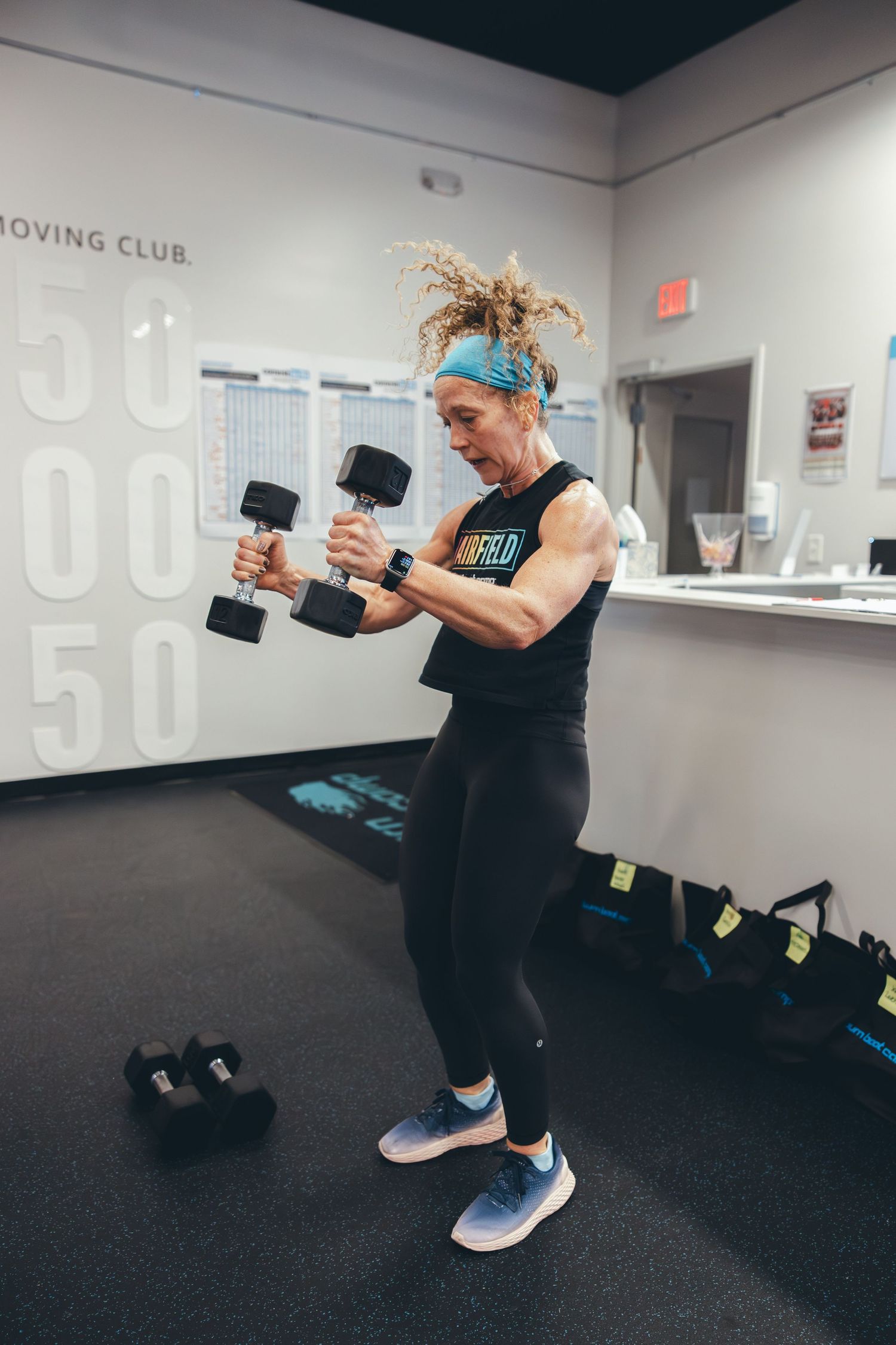 Fitness participant in black workout gear and blue headband performing dumbbell exercises in a training studio.