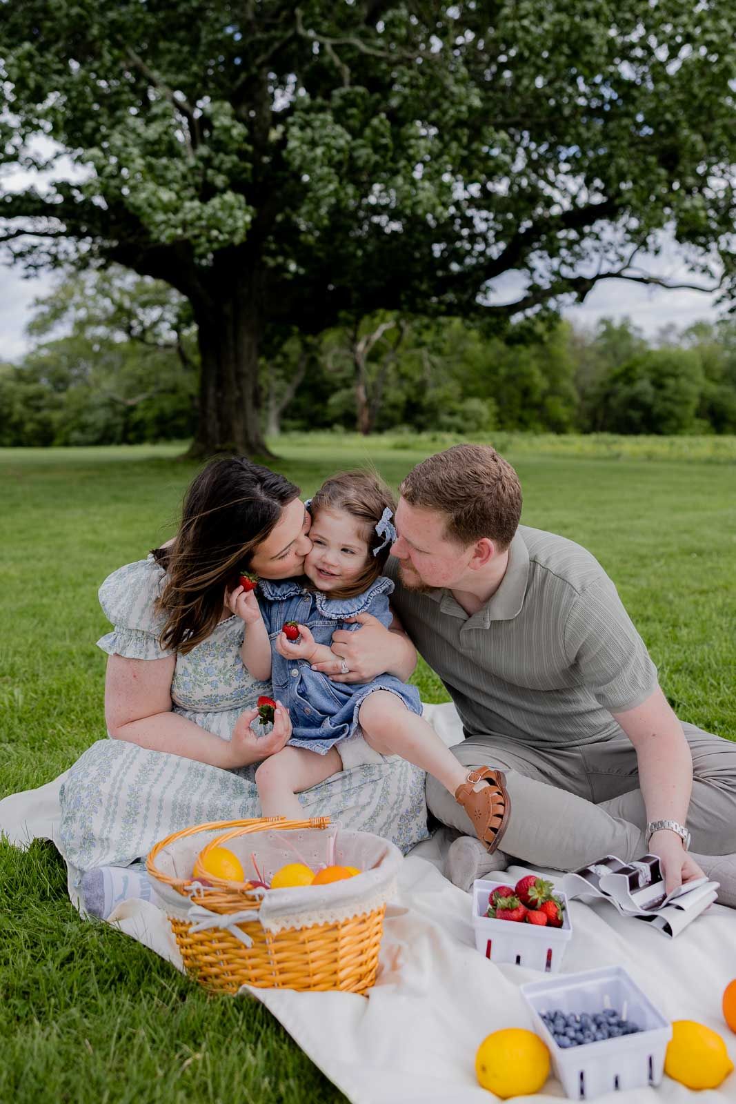 Family sharing intimate moment during picnic with fresh fruit and snacks on blanket.