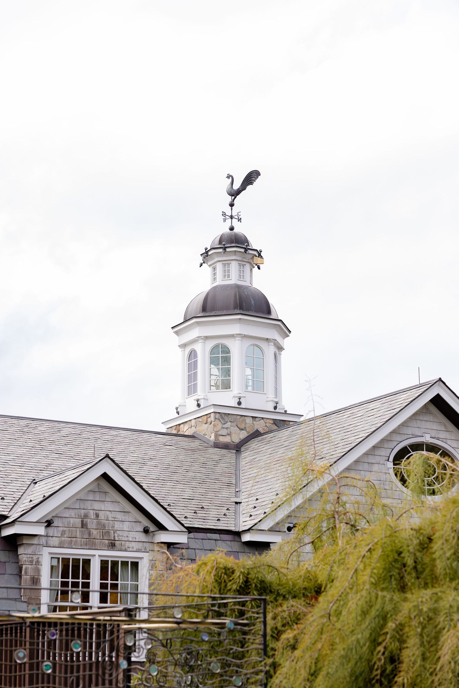 A classic white church steeple with a weathervane stands against a cloudy sky, framed by autumn trees.