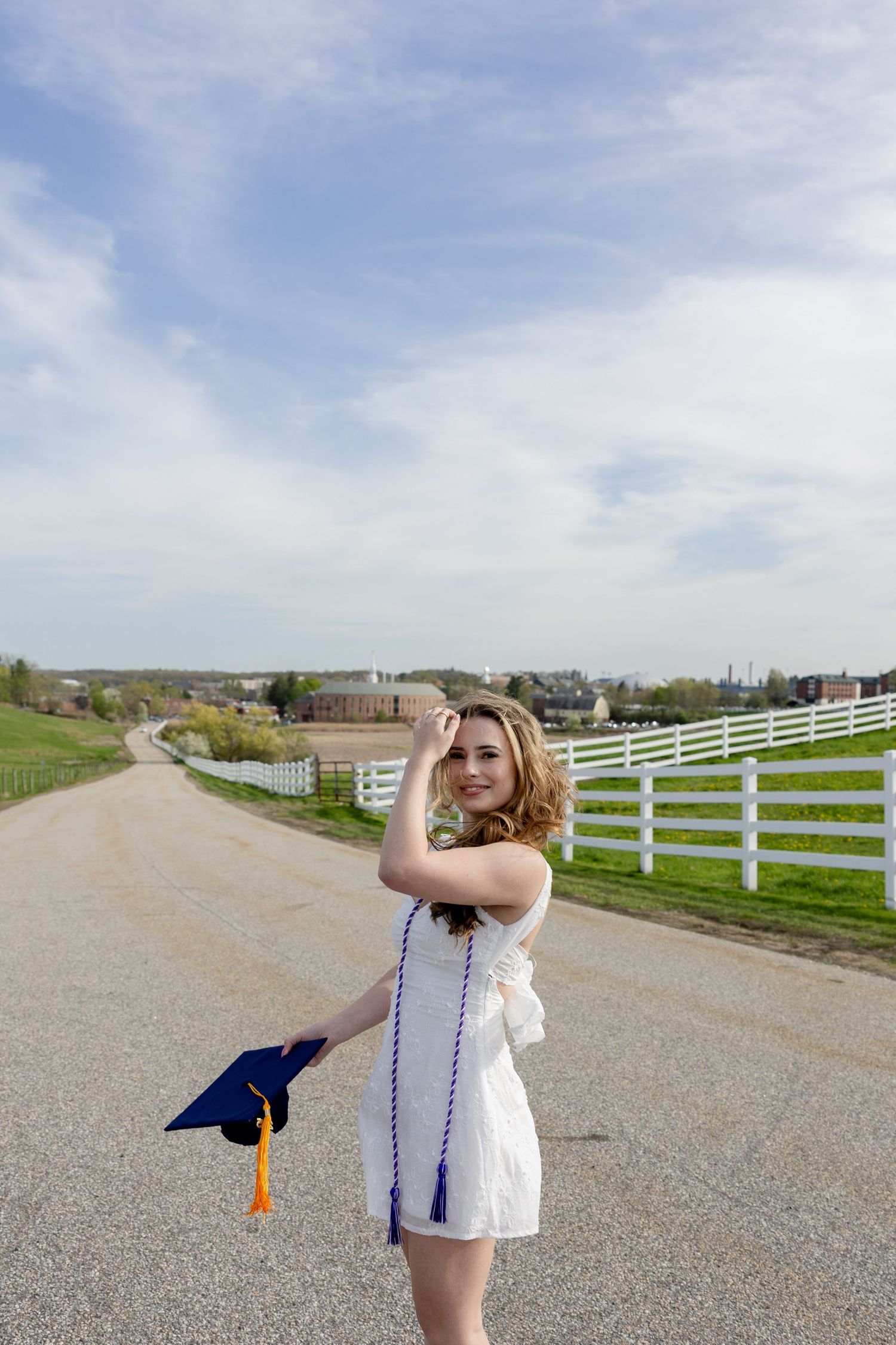 A person in a white dress stands on a rural road with white fencing and green fields, holding a blue graduation cap.