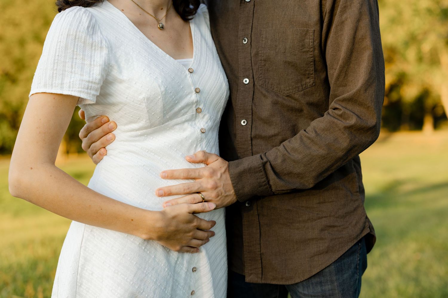 A couple stands close together in a sunny field wearing brown and white attire during a romantic photoshoot.