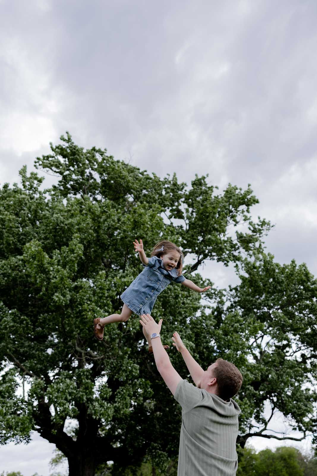 A playful moment as a parent tosses a small child high in the air against a backdrop of trees and cloudy sky.