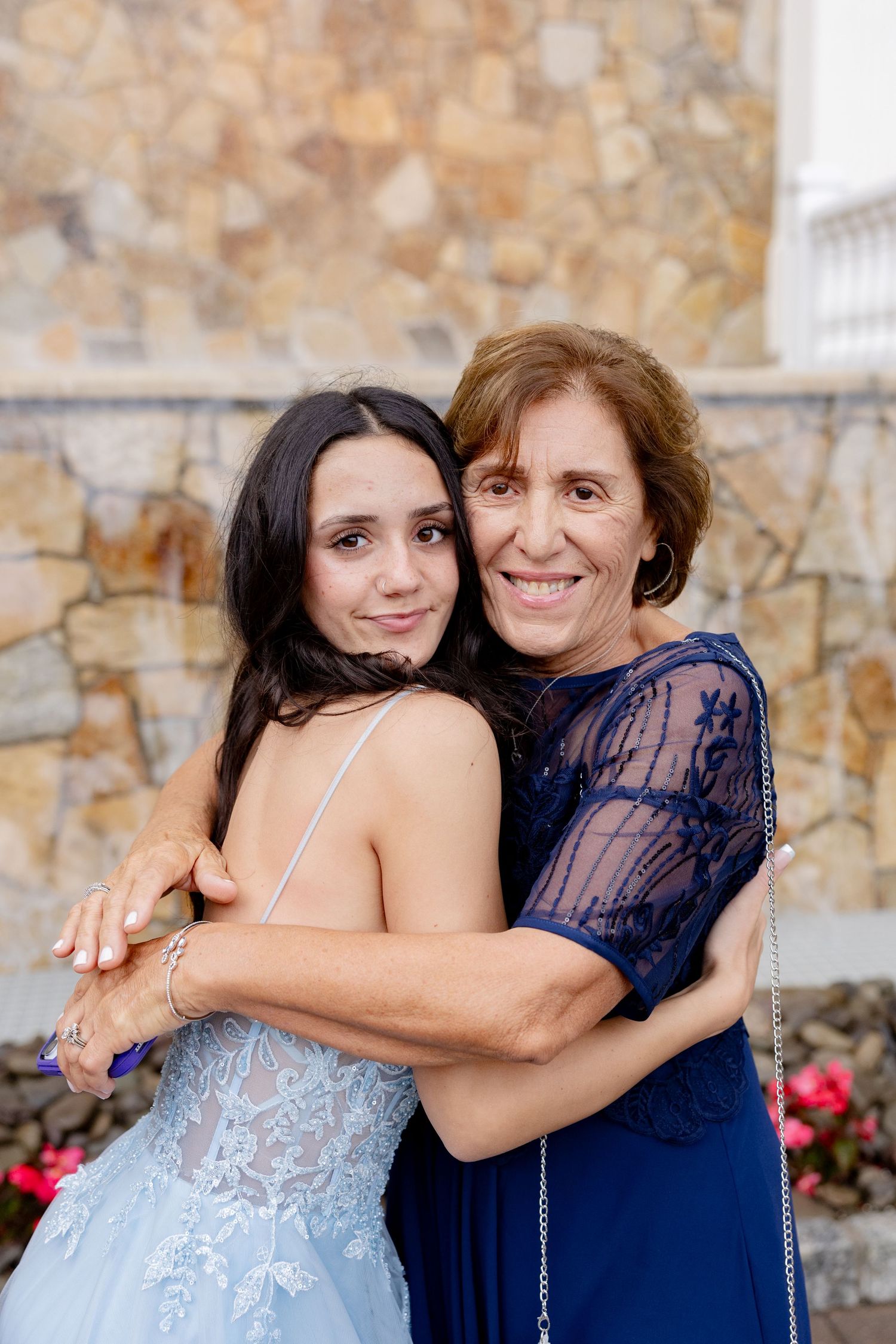 Two women in formal attire embrace while posing together against a stone wall with red flowers in the background.