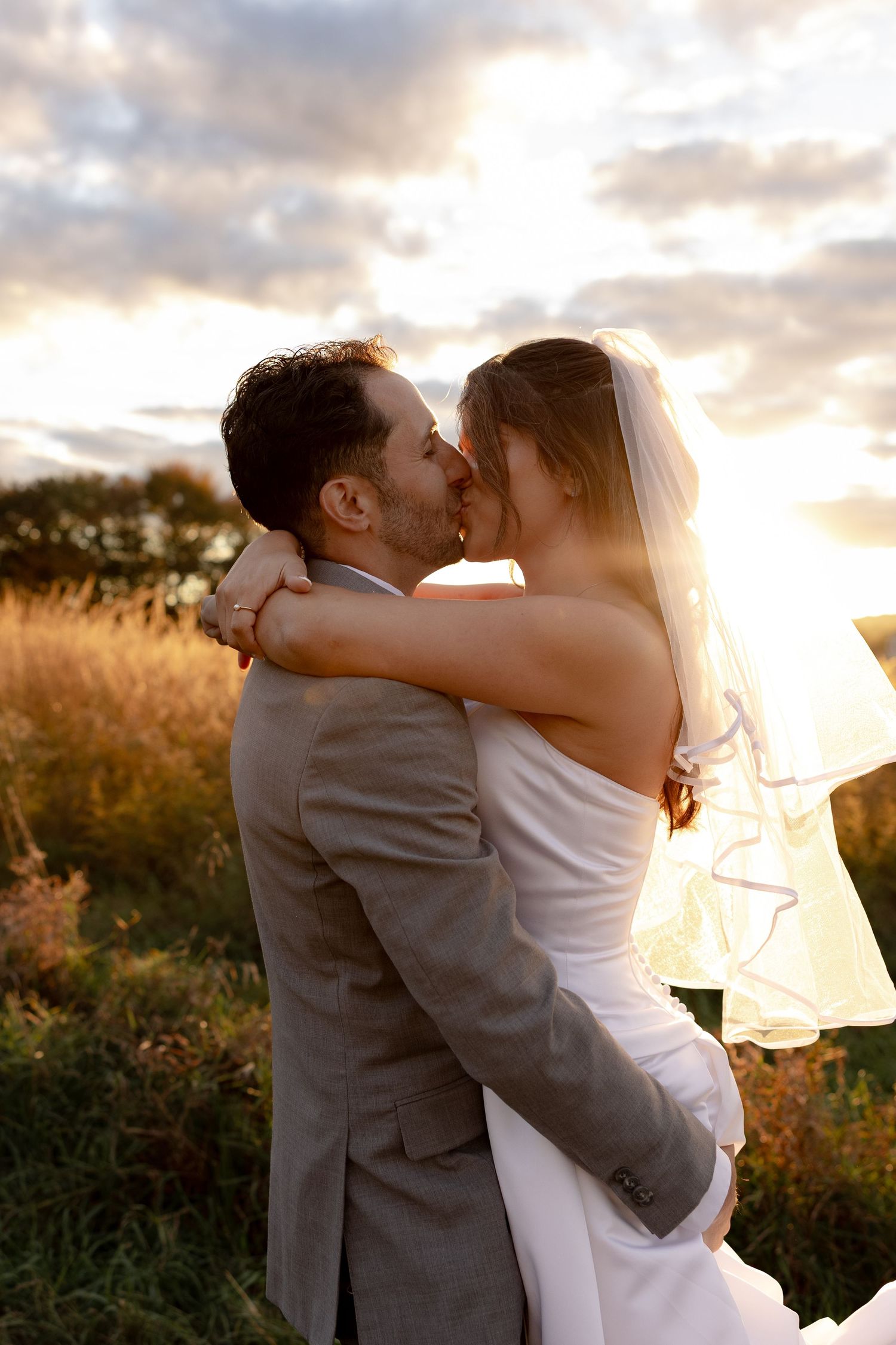 A romantic wedding couple embraces and kisses in a golden field at sunset.