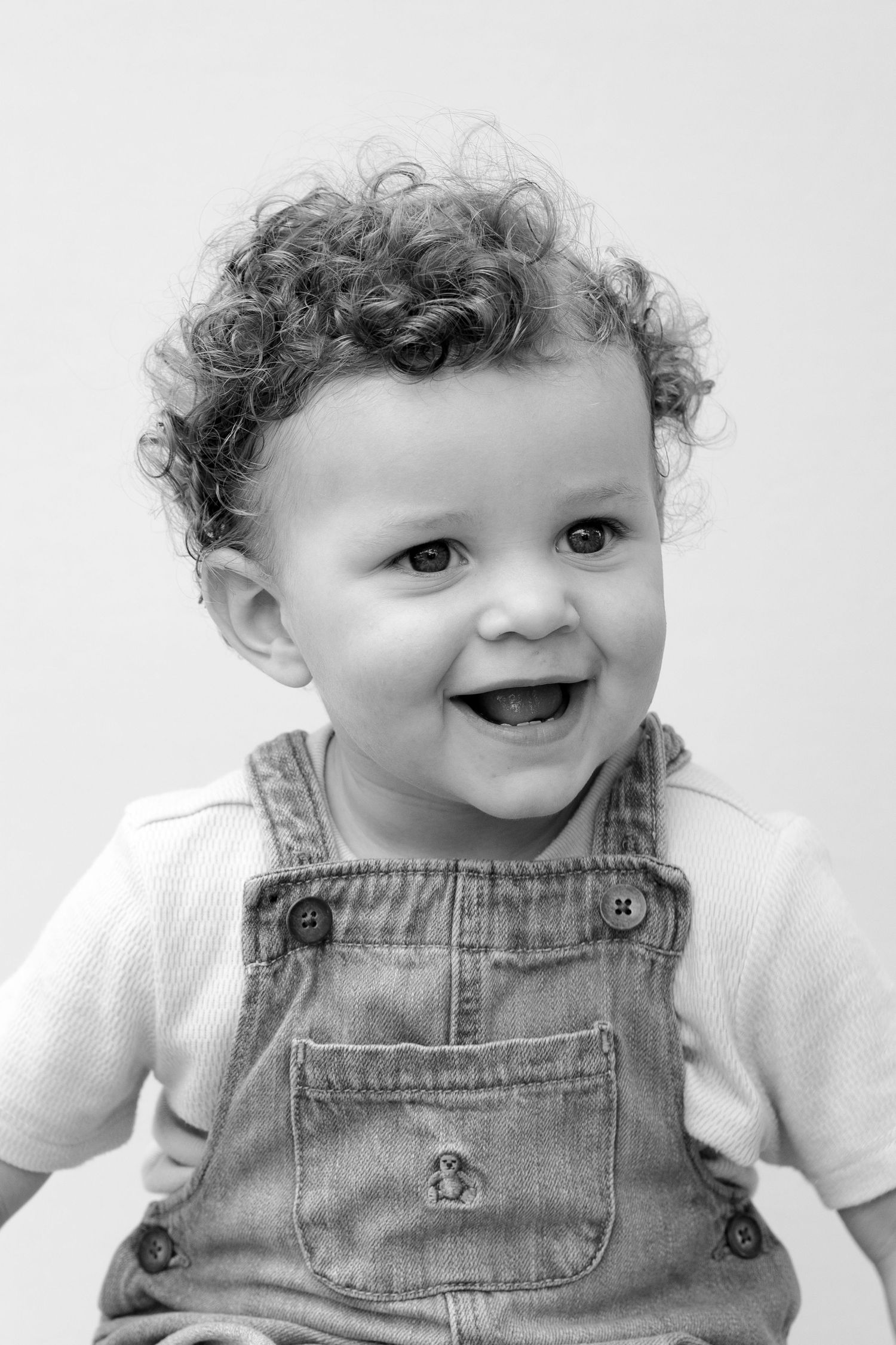 Black and white portrait of a happy toddler wearing denim overalls and a white shirt with curly hair.