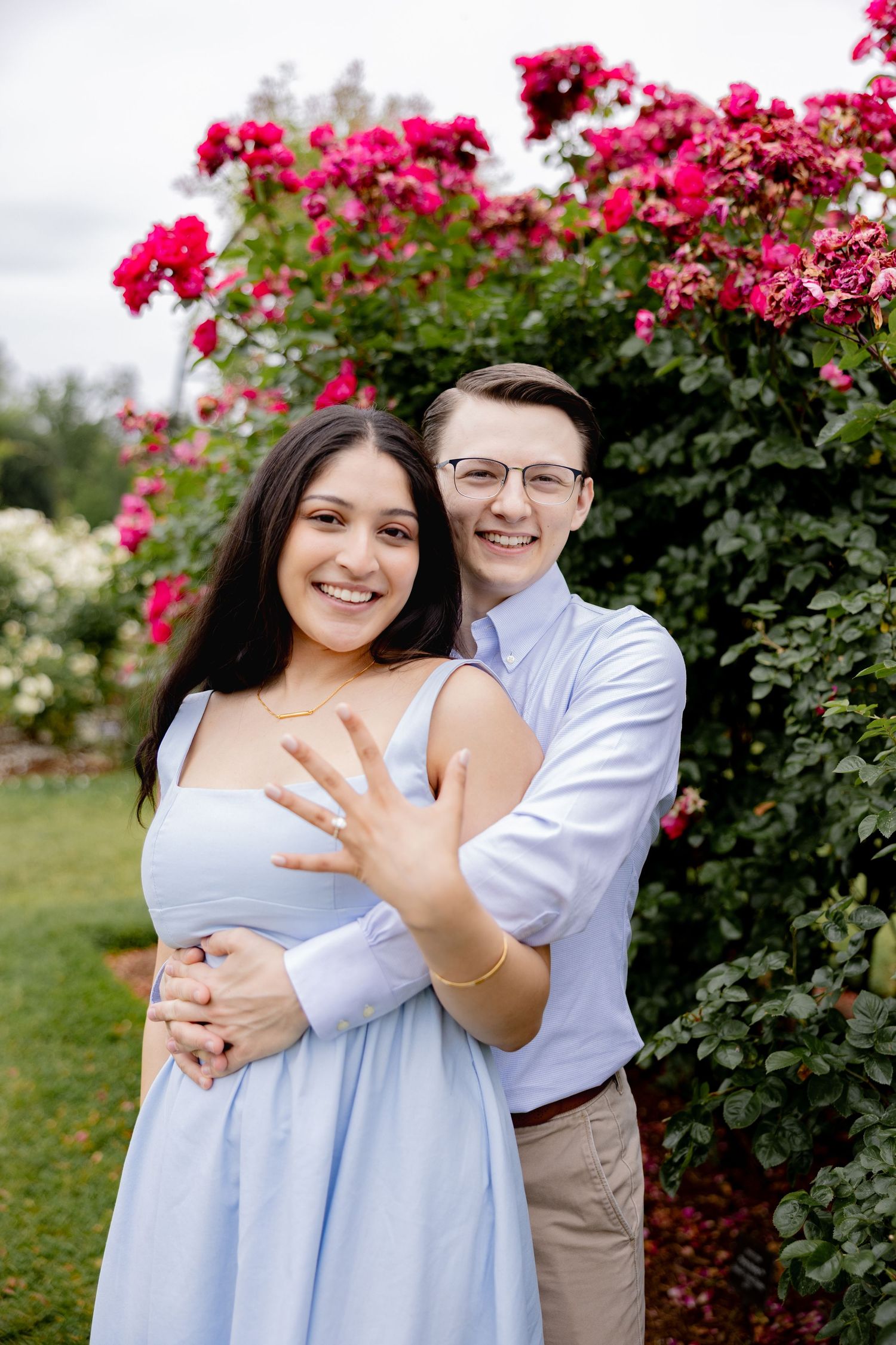 A happy couple embraces in a garden with pink roses as she shows off her engagement ring with a joyful smile.