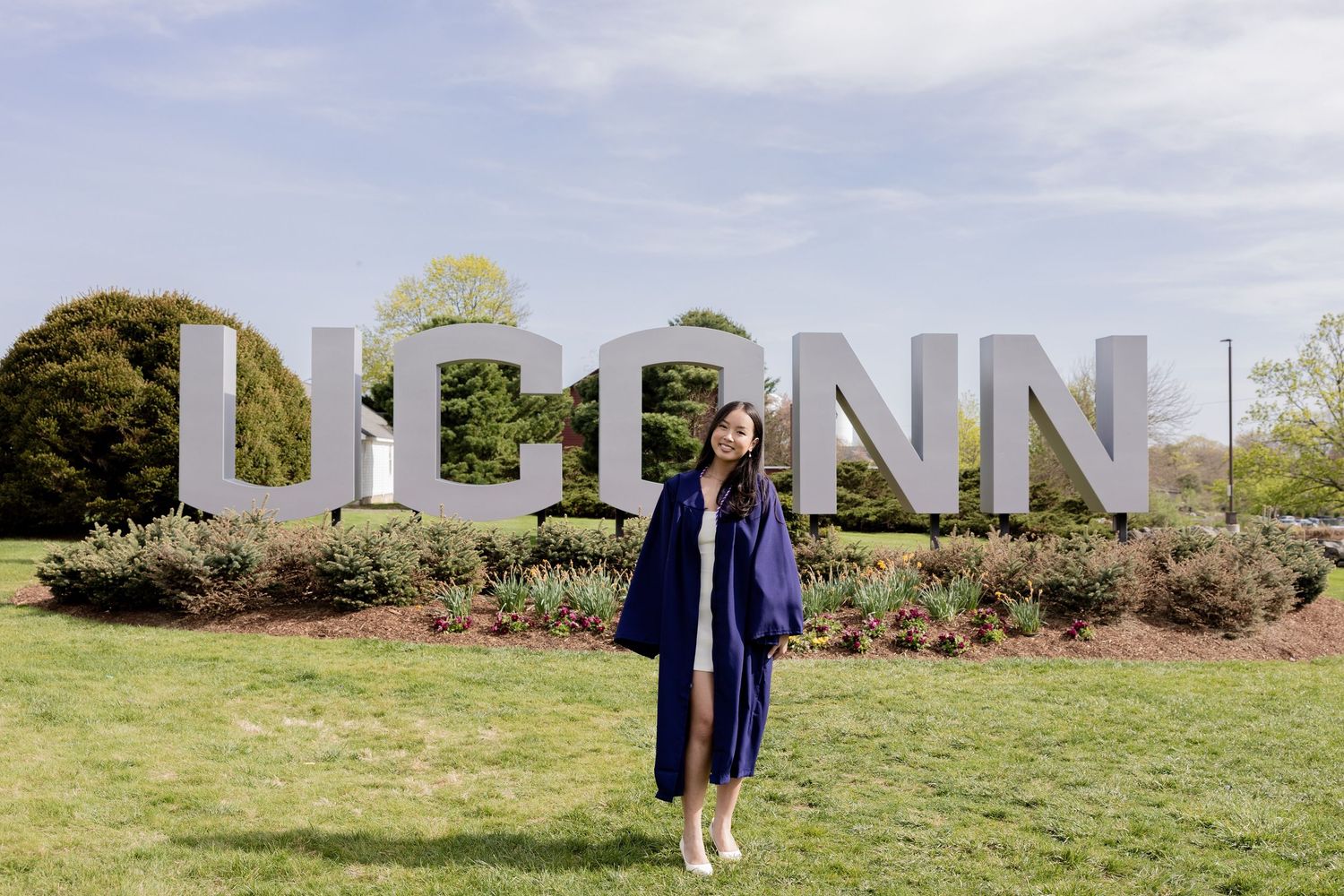 Graduate in cap and gown poses in front of large UPENN letters on a grassy lawn with trees and shrubs.