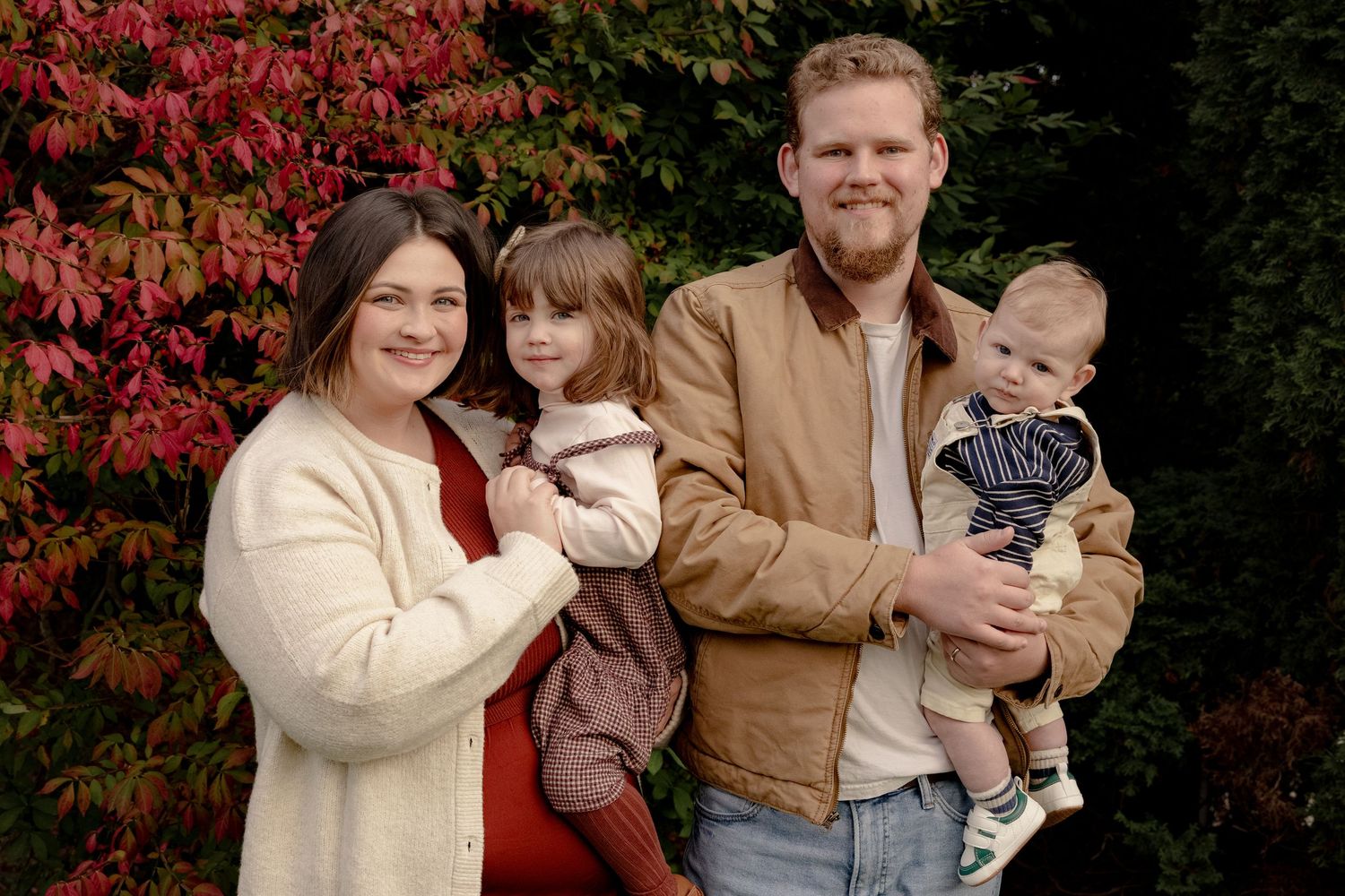A family poses for autumn portraits in front of vibrant red foliage wearing coordinated brown and cream outfits.