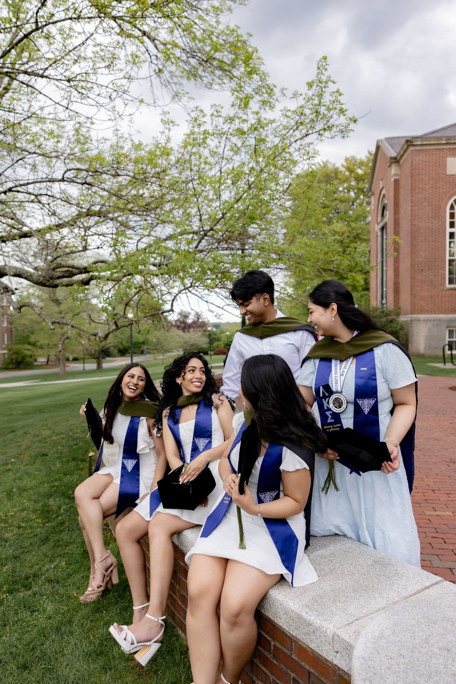 A group of graduates in blue and white stoles sit together on a brick ledge on a college campus during spring.
