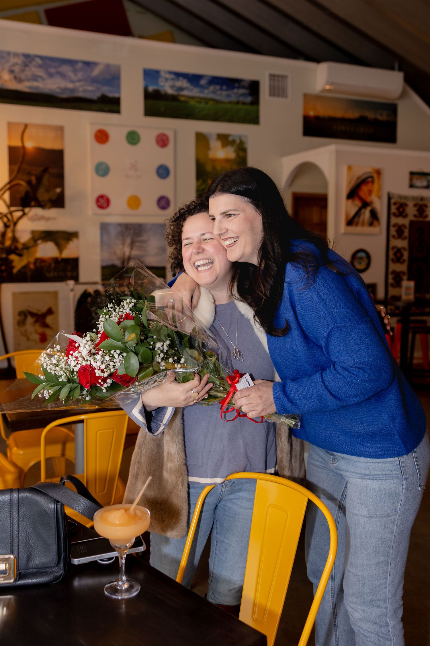 Two people laugh together while sitting at yellow chairs in a restaurant with floral decorations and artwork on walls.