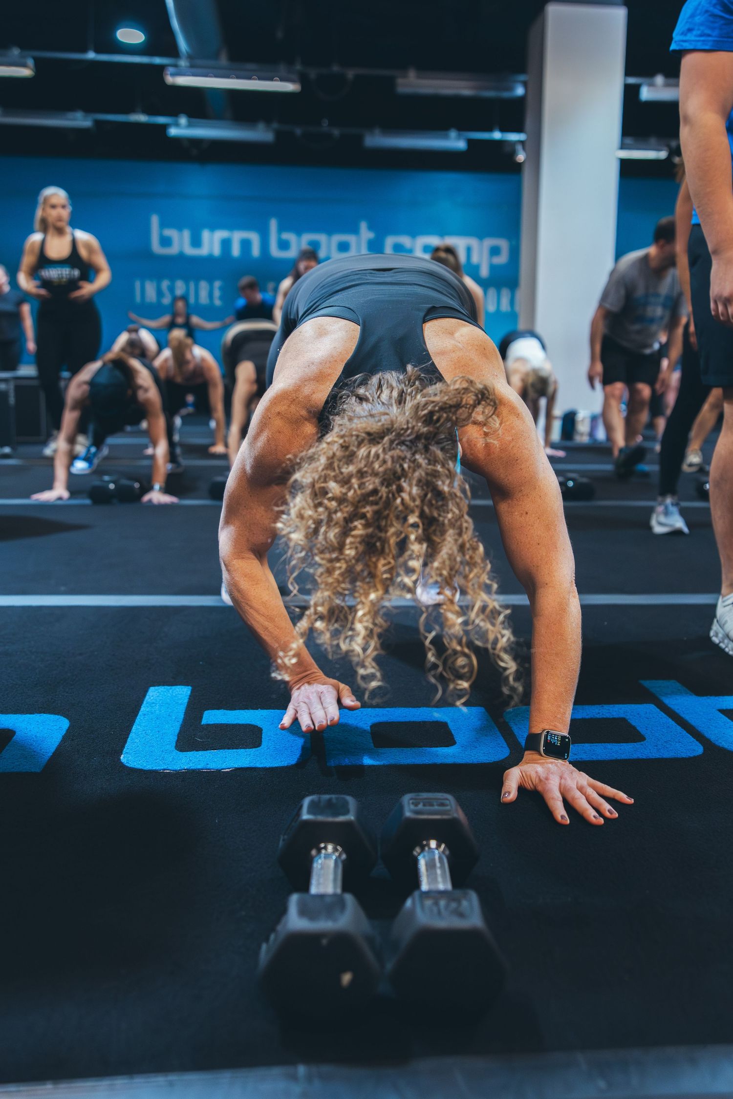 Athlete with curly hair performing a burpee exercise on blue gym floor with weights nearby.