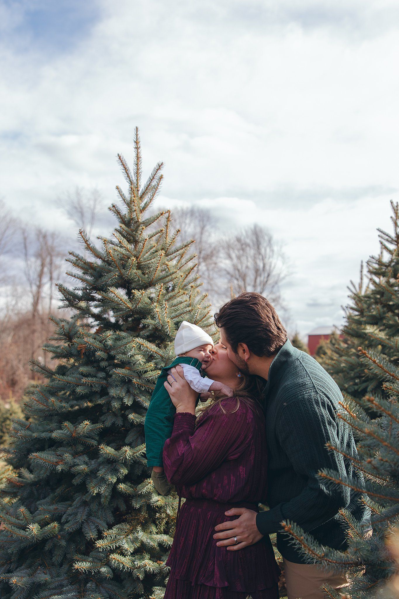 mom and dad kissing baby in front of pine trees on maple row farm
