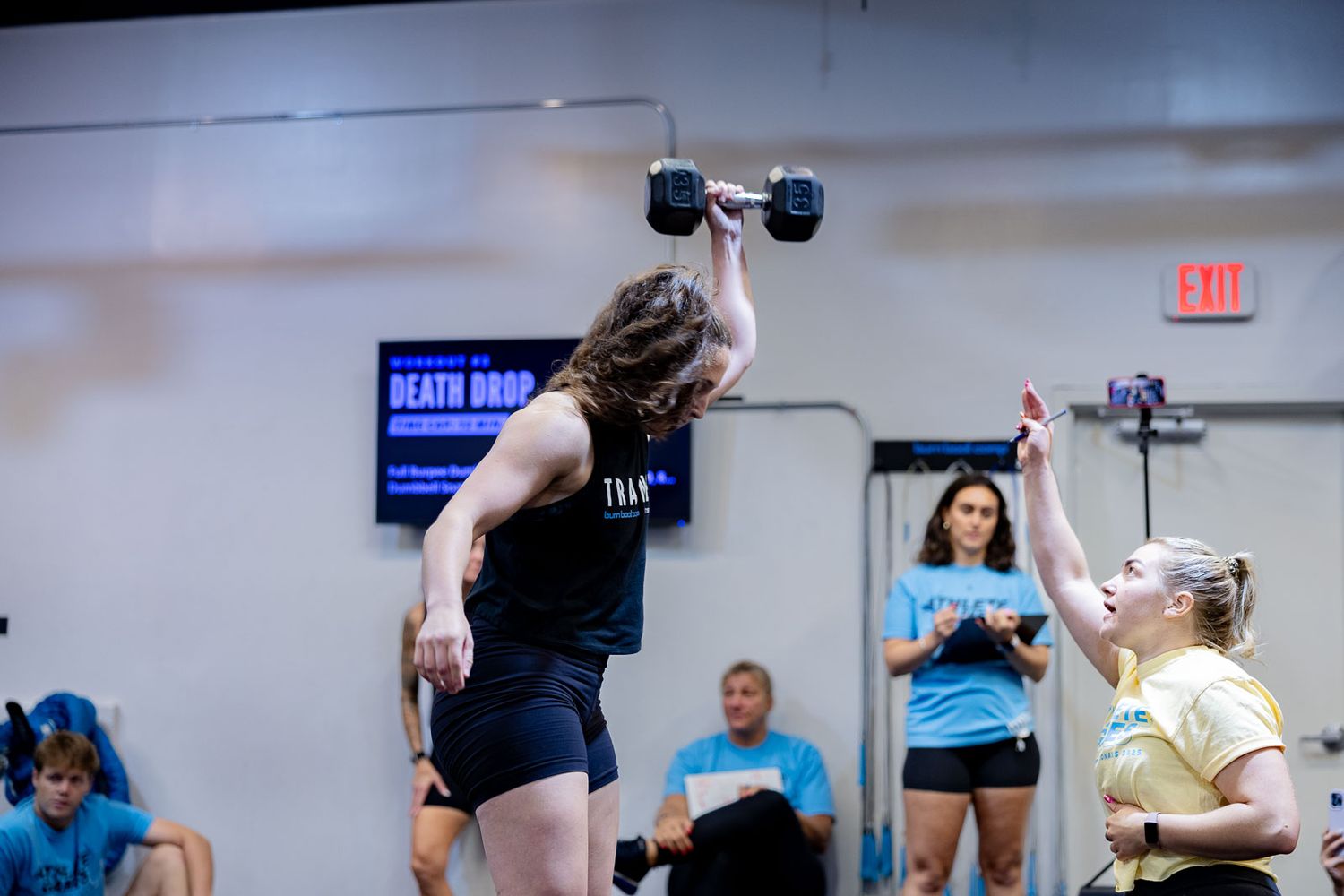 An athlete lifting dumbbells overhead during an indoor weightlifting competition or training session.