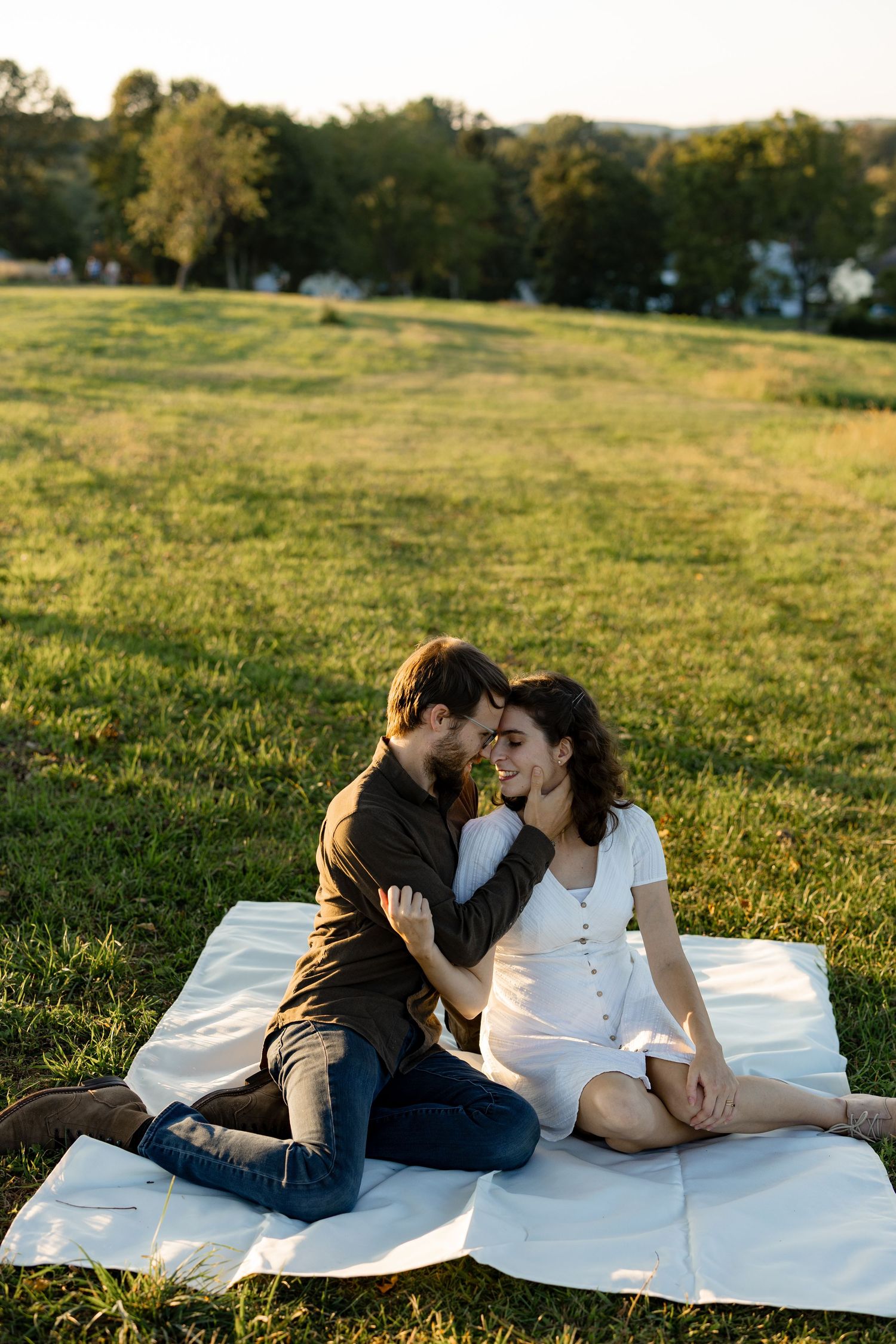A couple shares an intimate moment on a white blanket in a sunlit grassy field during golden hour.