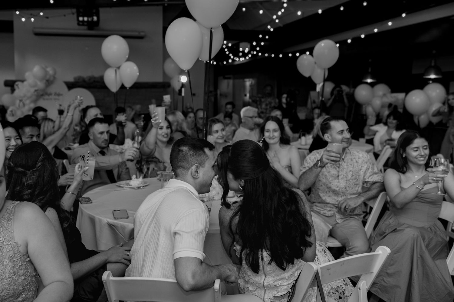Wedding reception guests dance and celebrate under white balloons and string lights in an indoor venue.
