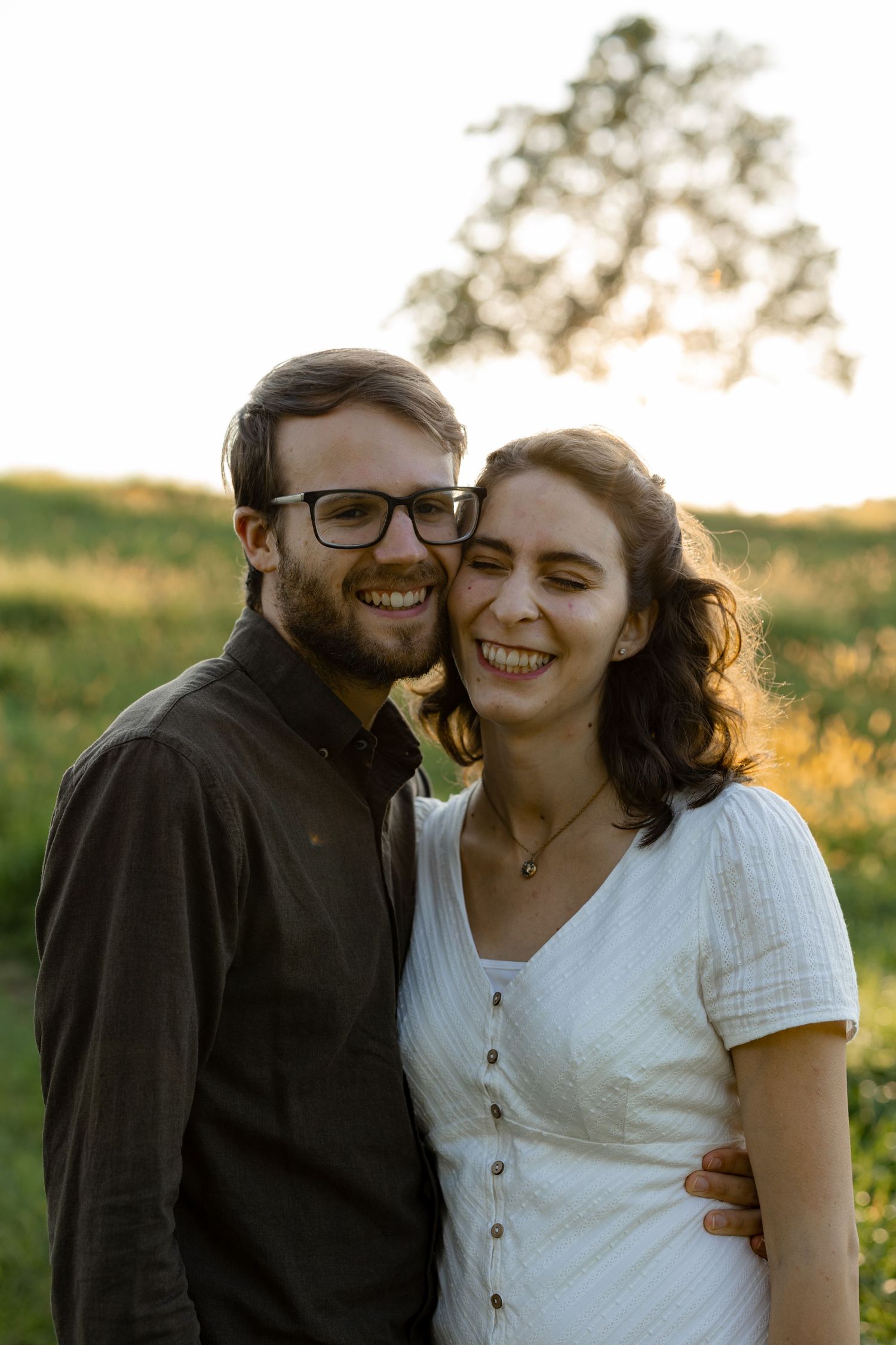 A happy couple embraces and smiles together in a grassy field at sunset.
