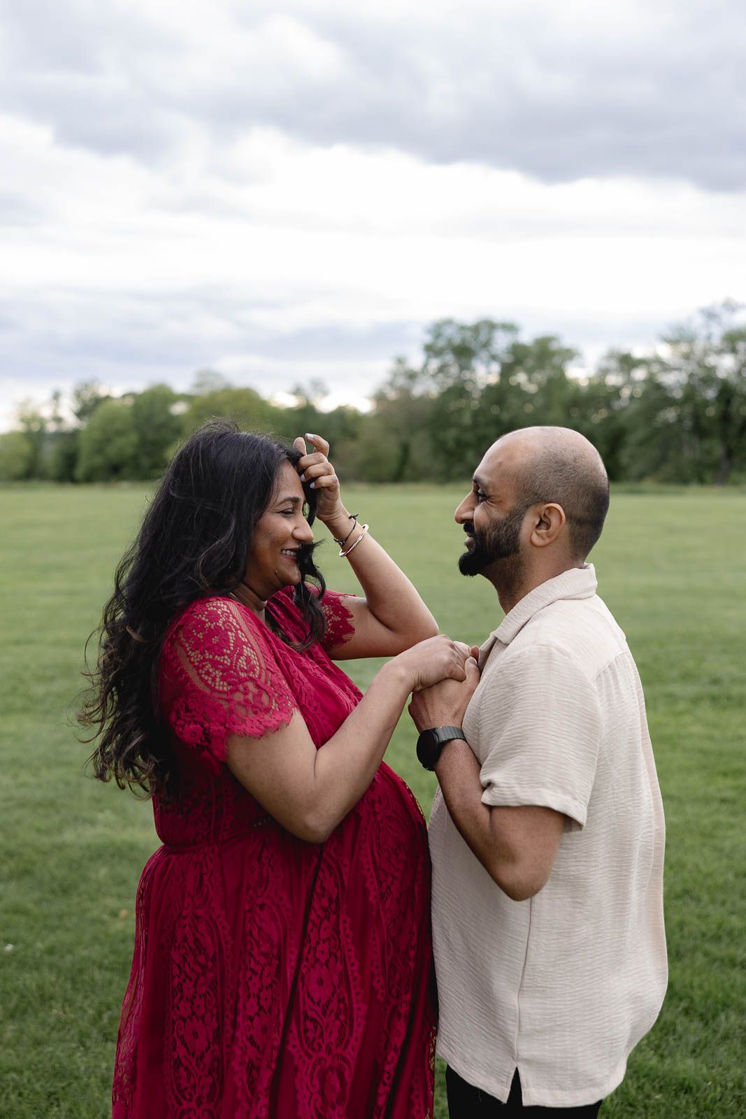 A playful couple laughs together in a green field, the woman wearing a red dress and man in beige shirt.