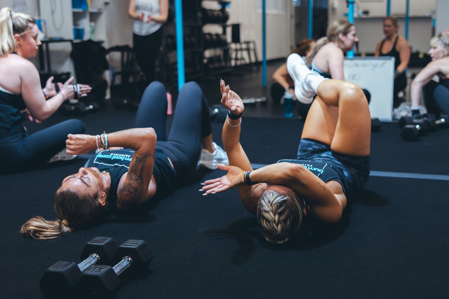 Athletes lying on gym floor doing core exercises with dumbbells during group workout.