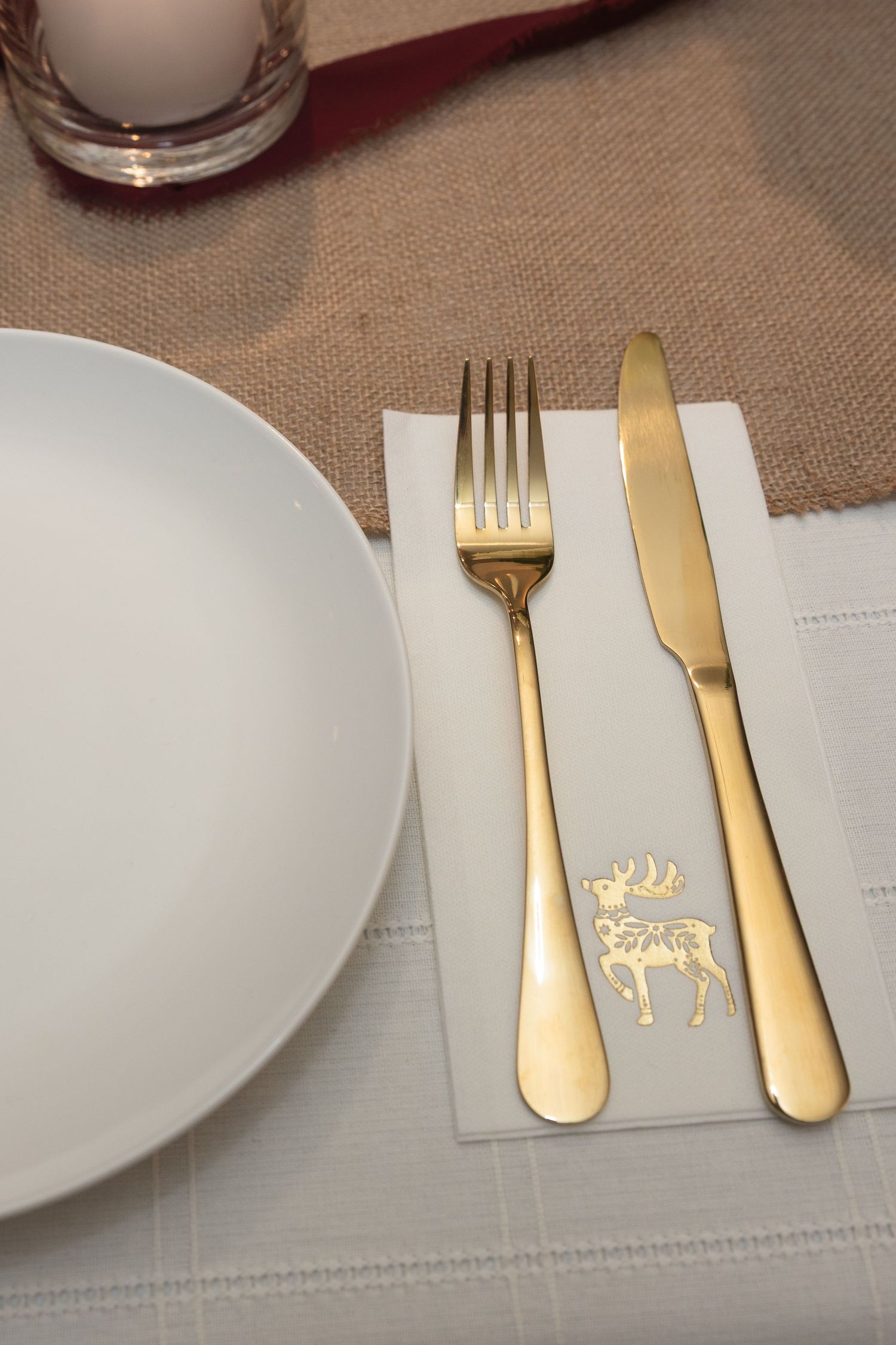 Place settings at a formal dinner table with gold utensils and white plates on a beige linen tablecloth.
