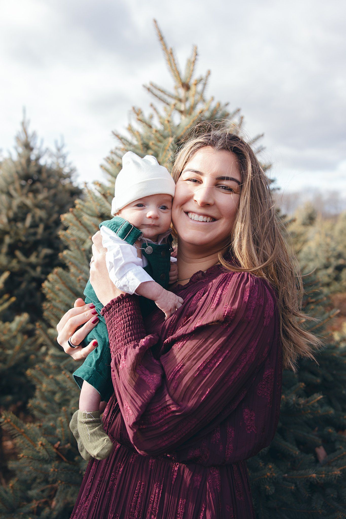Mother in burgundy dress holds baby against backdrop of evergreen trees.