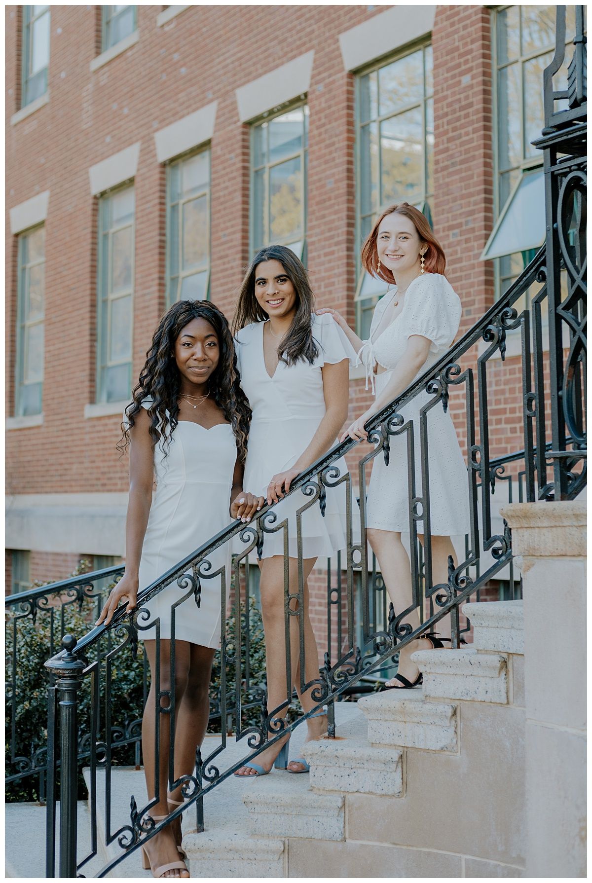 Three friends in white dresses pose on historic brownstone steps with ornate iron railings.