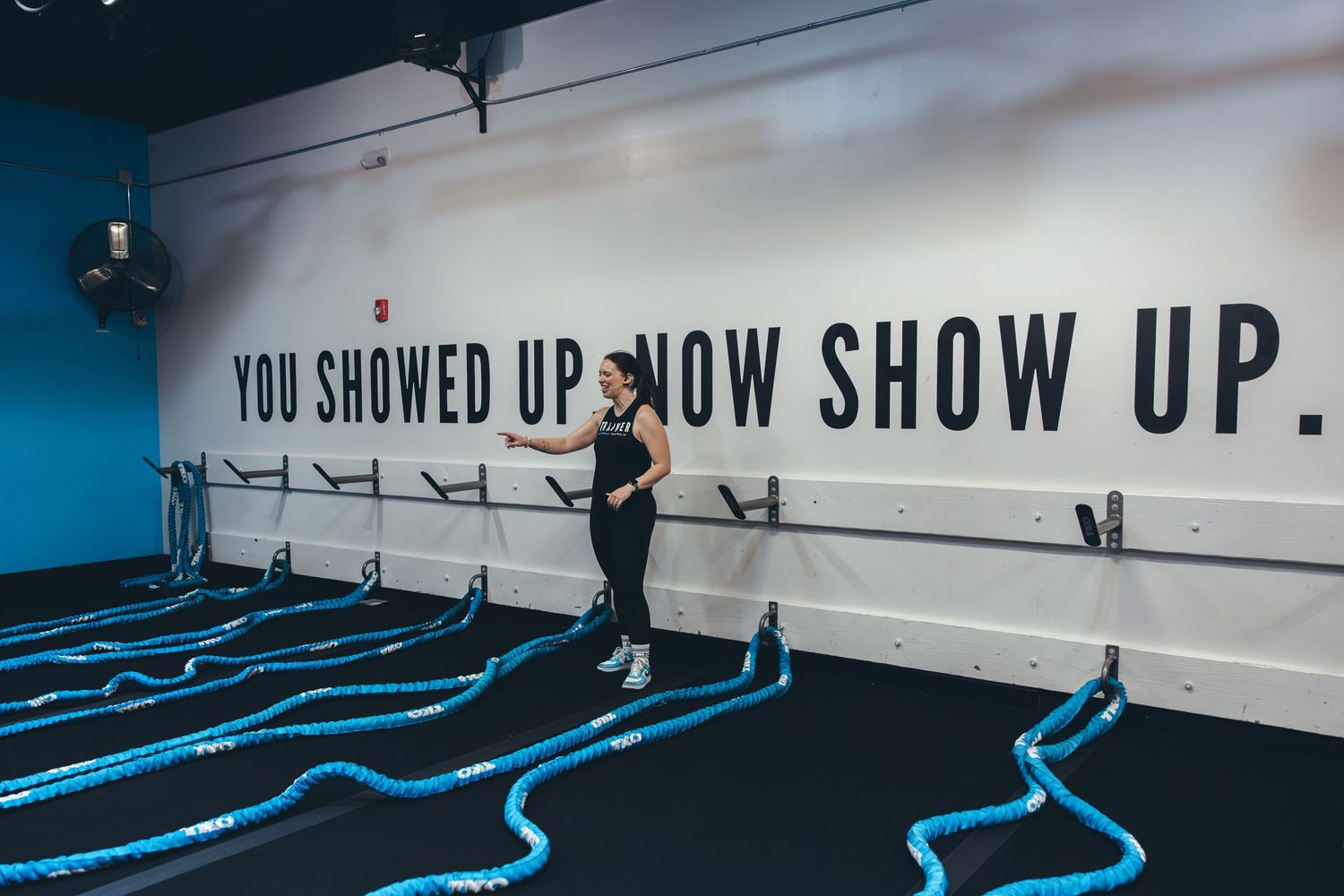 Person exercising in a modern gym with blue floor lines and motivational text 'YOU SHOWED UP NOW SHOW UP' on white wall.