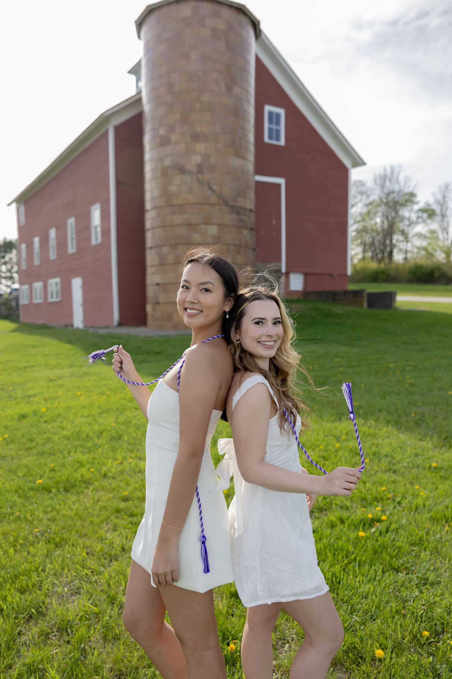 Two friends in white summer dresses posing playfully with bubble wands in front of a red barn on a sunny day.