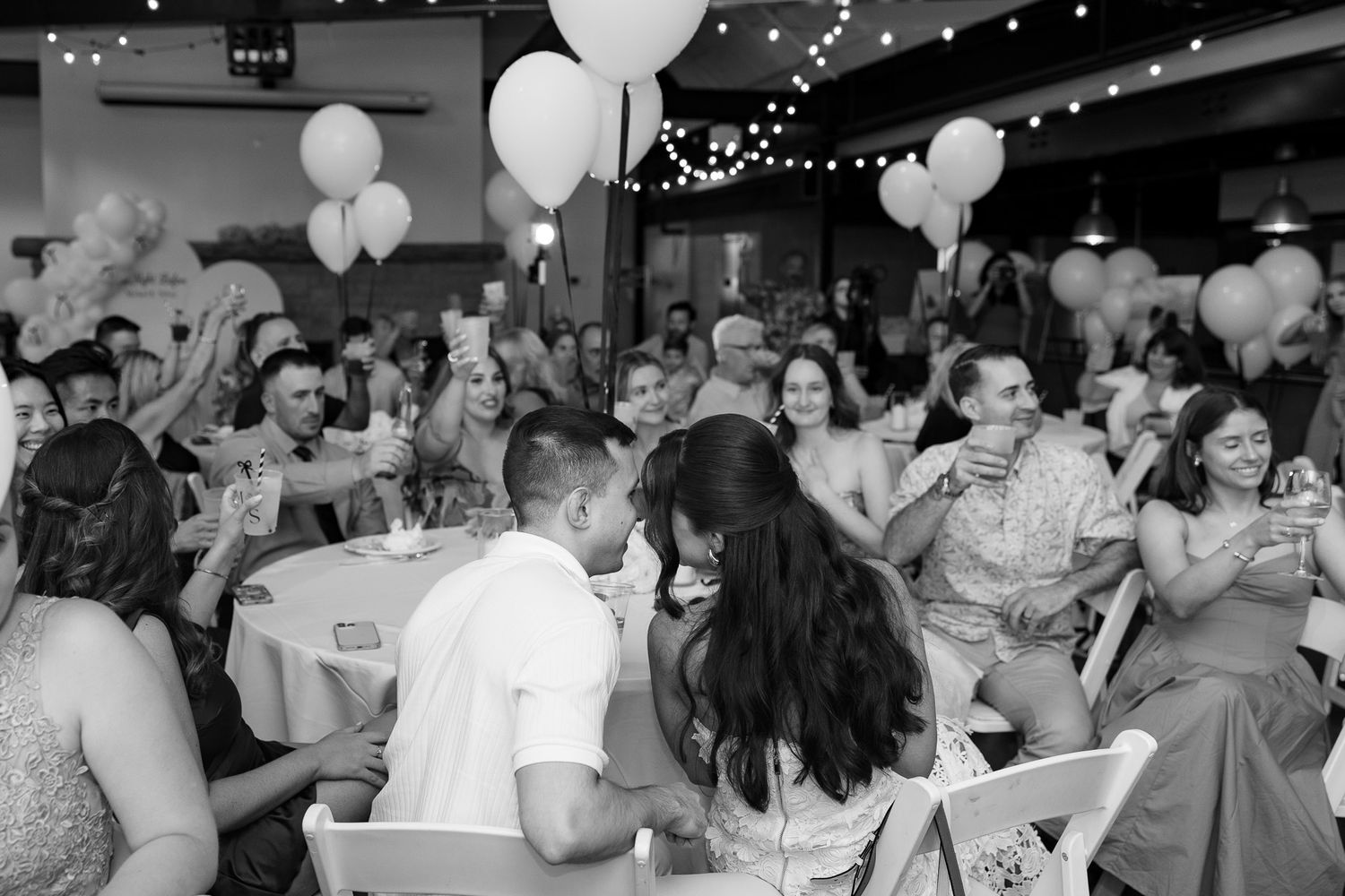 Wedding reception guests dancing and celebrating under white balloons and string lights.