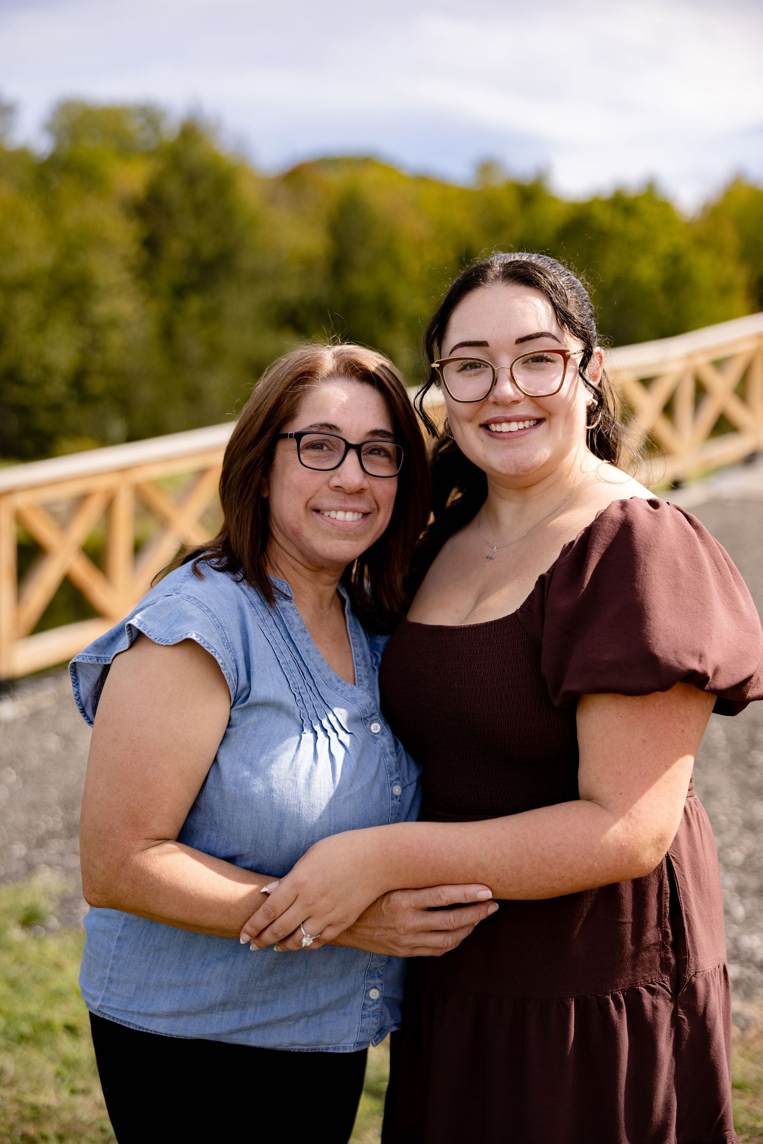 Two people wearing casual clothing embrace and smile while standing outdoors in a natural setting with wooden railings.