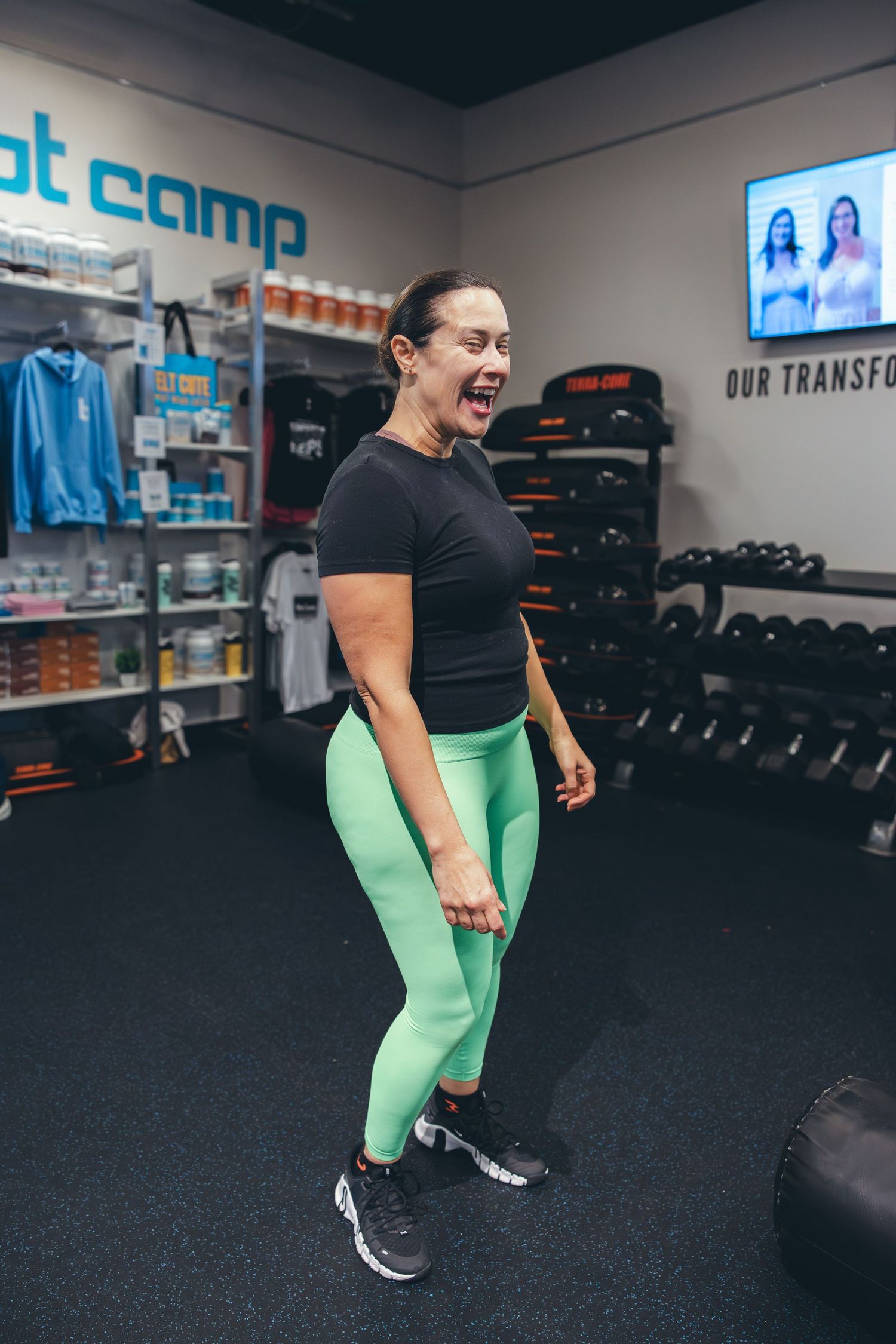 Woman in black top and mint green leggings exercising in a gym with dumbbell racks in the background.
