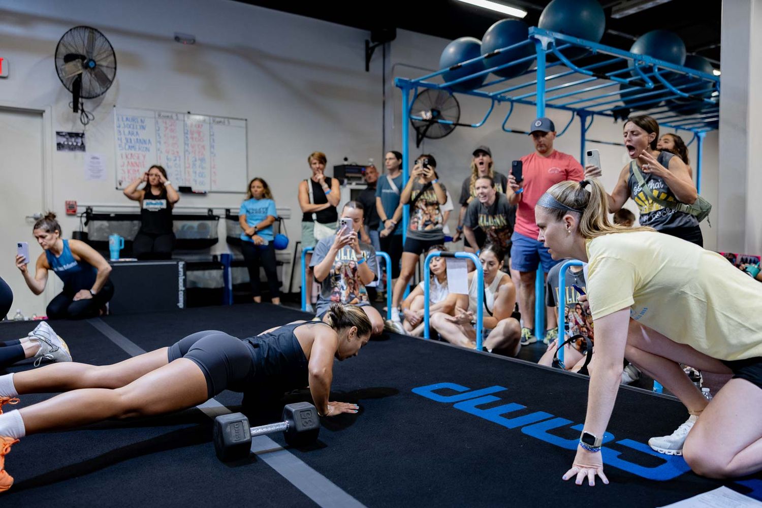 Group fitness class participants watching demonstration of proper plank form on black exercise mat in gym studio.
