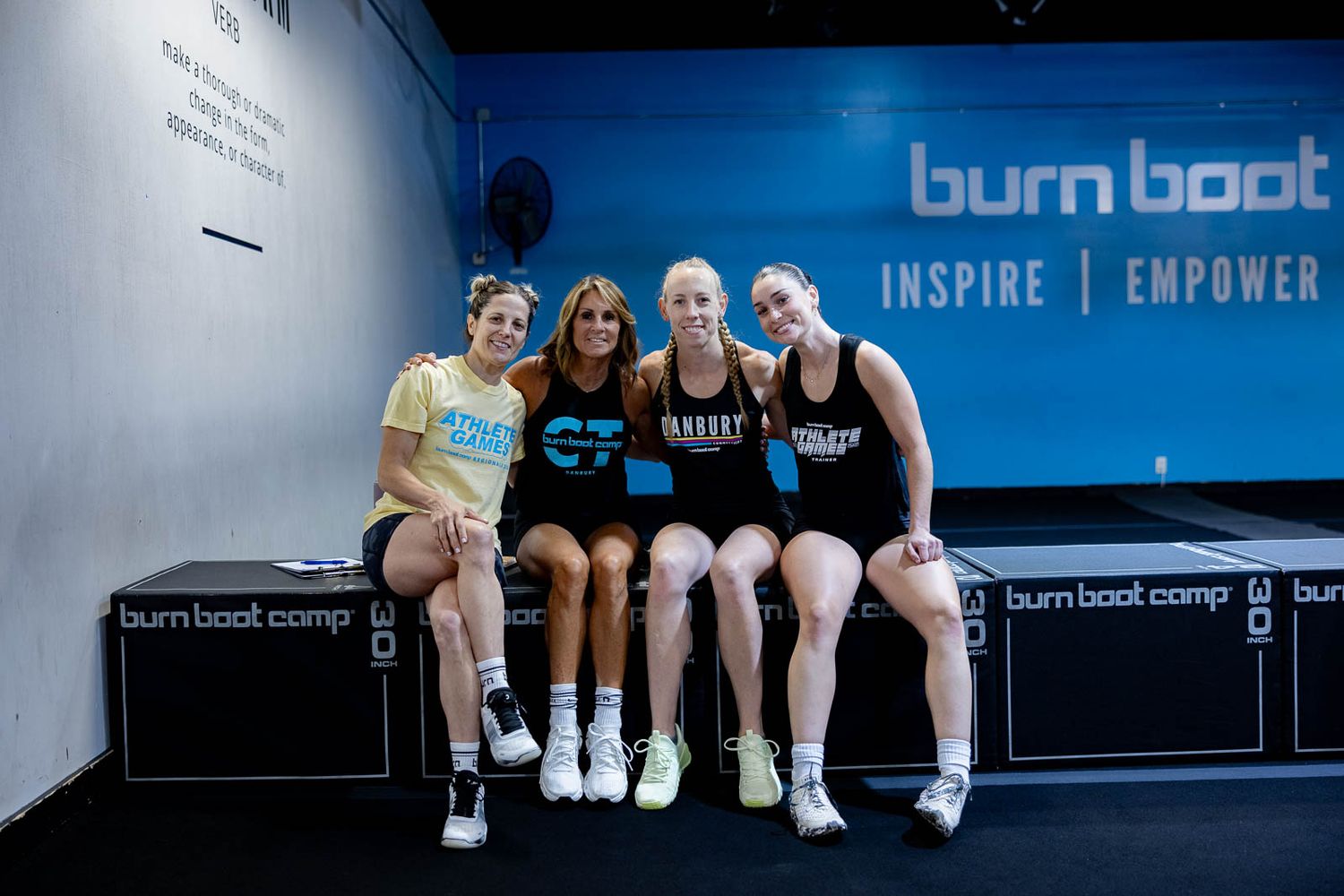 Four people sit together smiling on a bench in front of a blue wall with 'burn boot' signage.