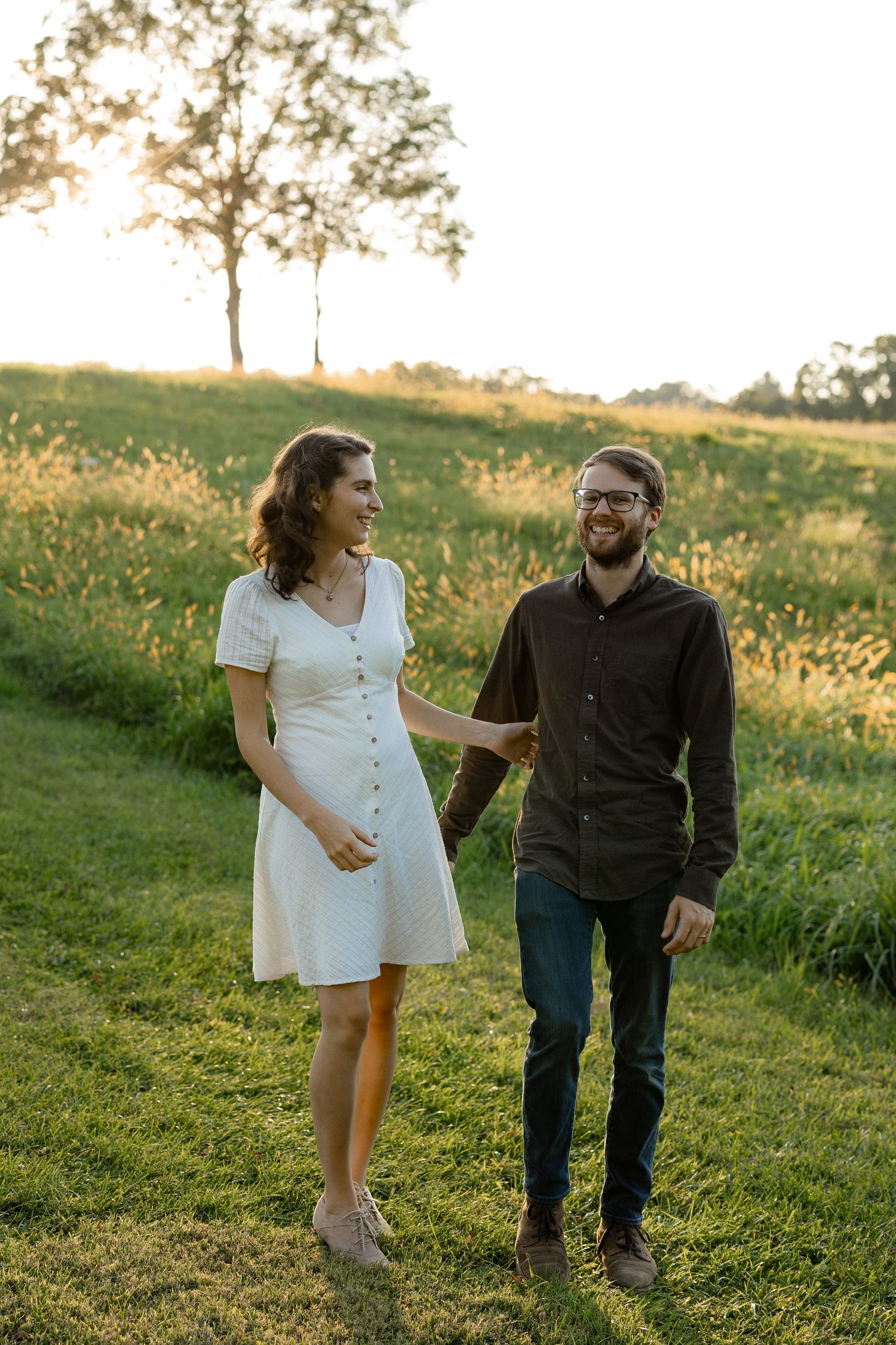 A couple laughs together while walking through a sunlit meadow at golden hour.
