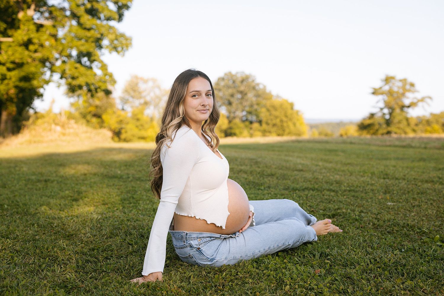 Expectant mother in white top and jeans sits on grass in golden sunlight.