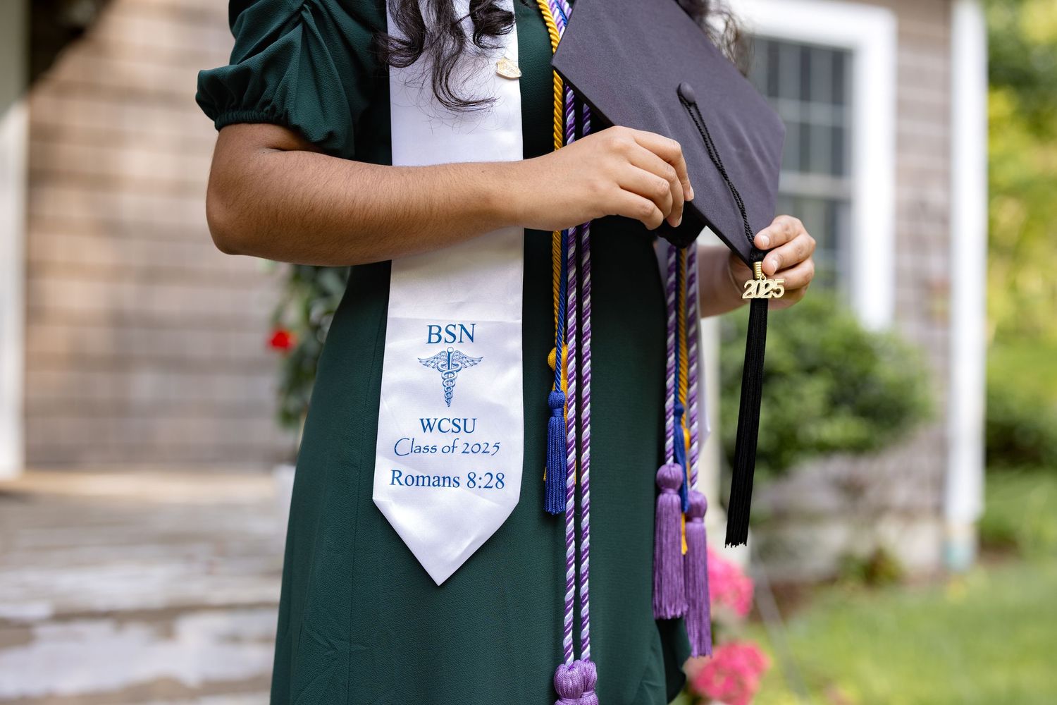 A person wearing a green graduation gown and white tie stands outside during a graduation ceremony.