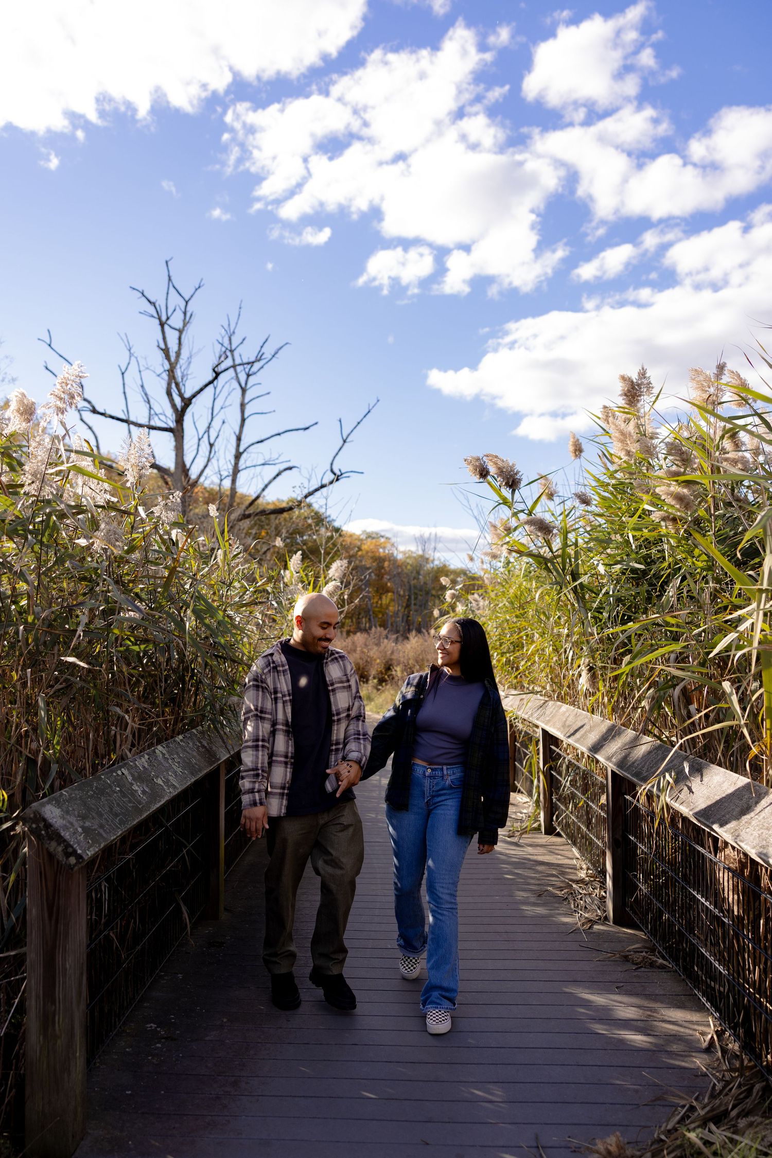 Two people walk together on a wooden boardwalk path surrounded by tall reeds and grasses on a sunny autumn day.