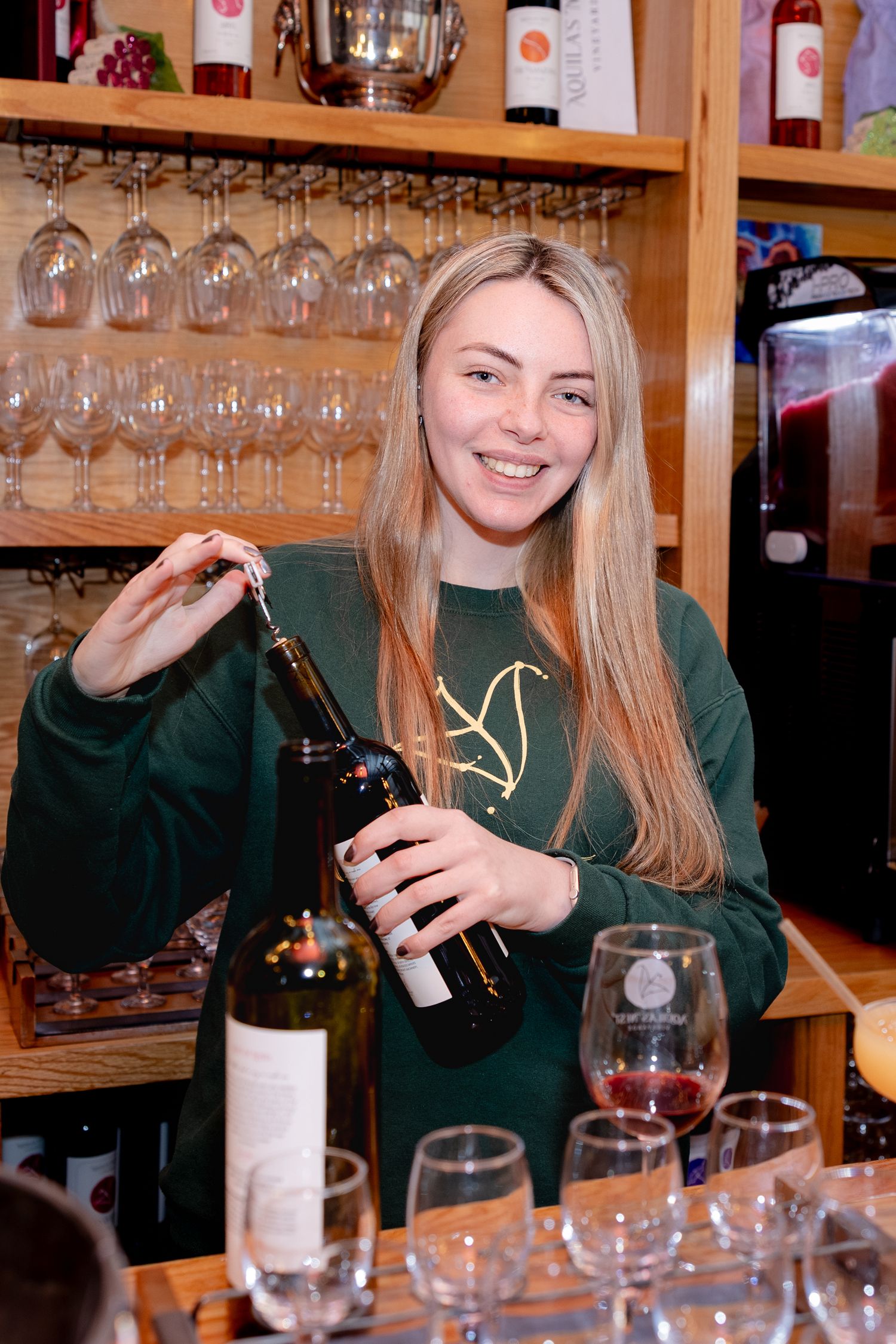 A person in a green sweater opening a wine bottle behind a bar counter with wine glasses displayed on shelves.