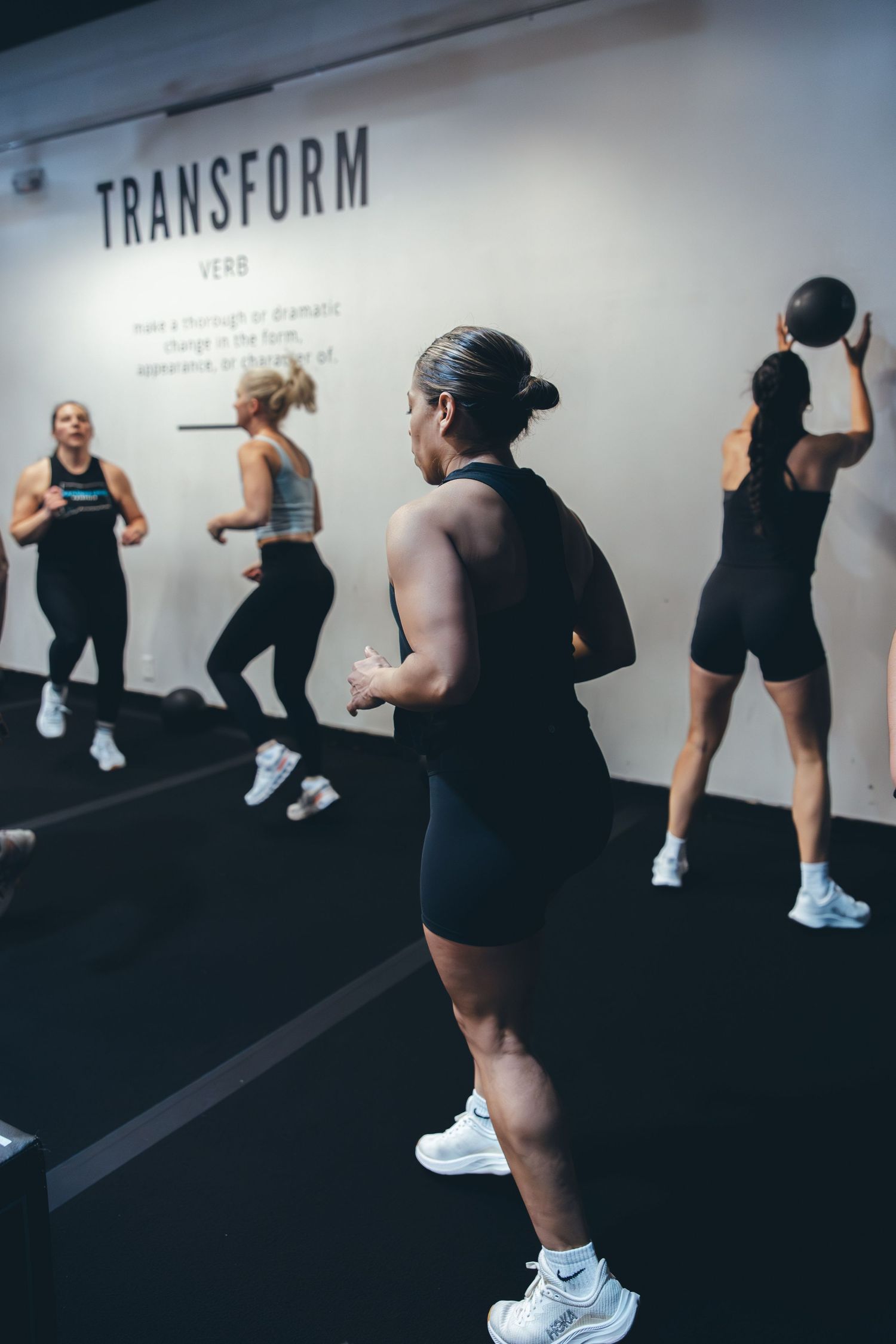 Group fitness class participants exercise with medicine balls in front of a 'Transform' wall sign.