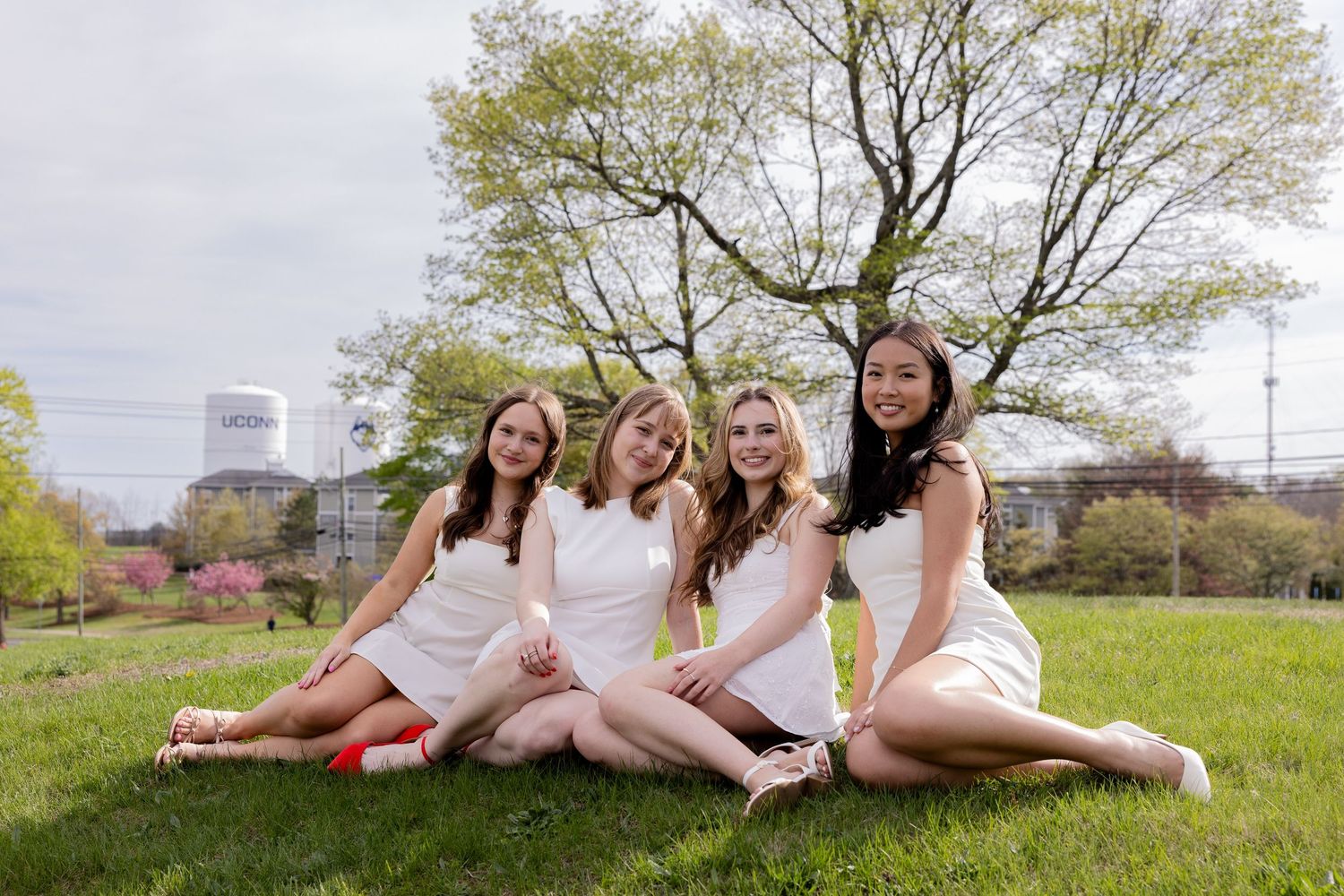 Four figures in white dresses pose together on green grass with spring trees and buildings in the background.