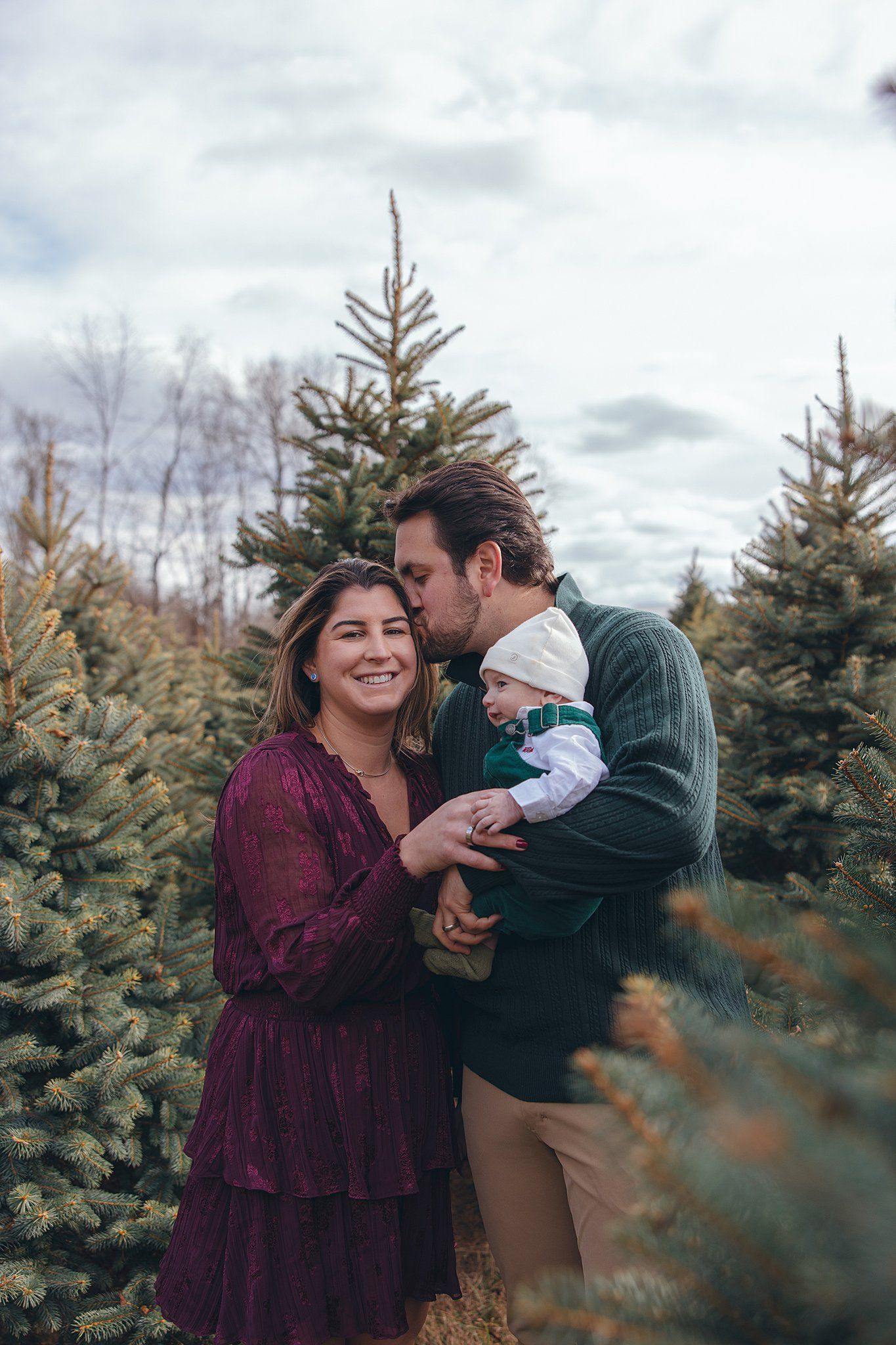 mom and baby smiling at camera and dad kissing mom on the cheek in front of a pine tree on maple row farm for holiday photo