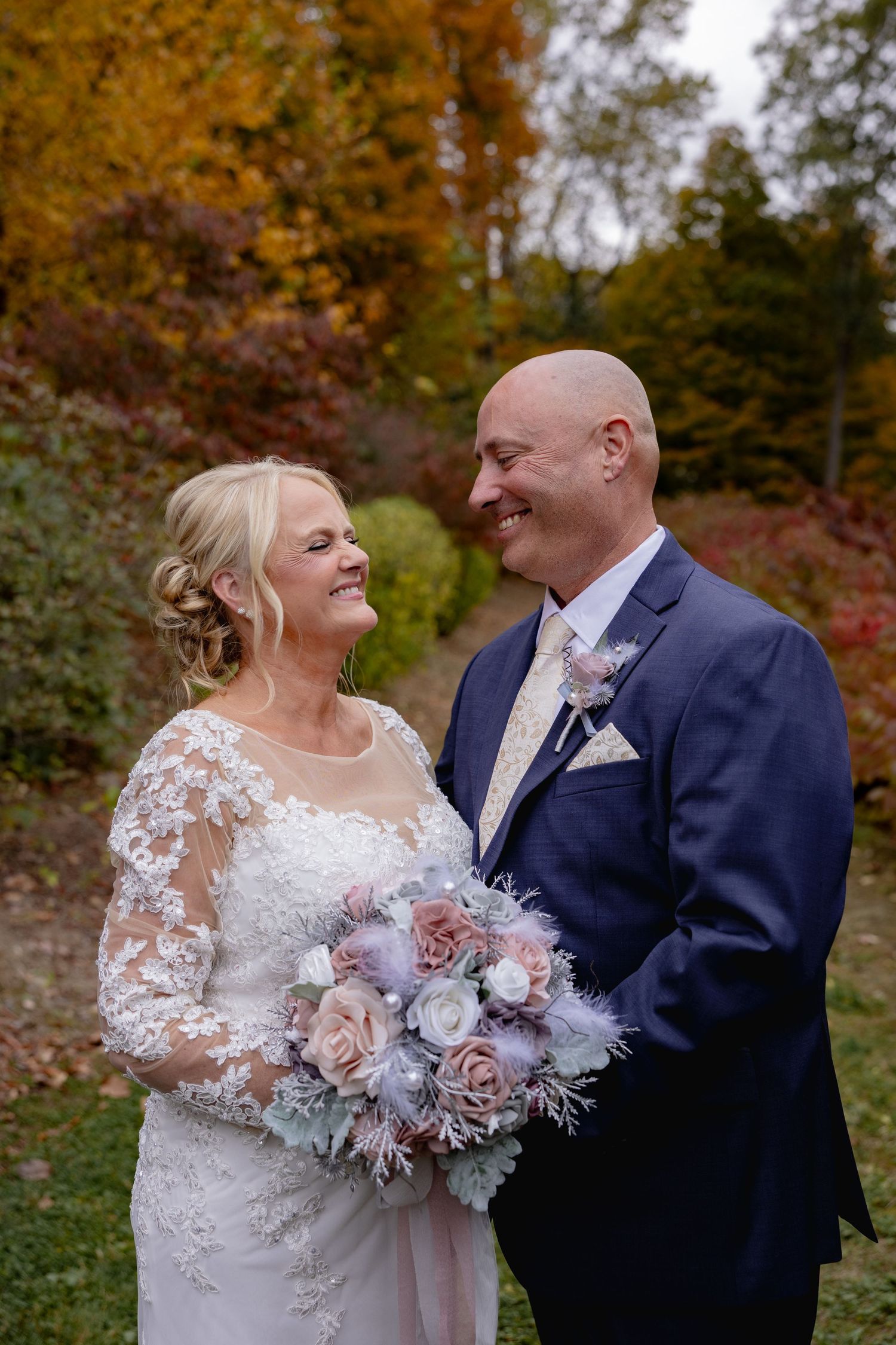 A happy couple shares a joyful moment surrounded by autumn foliage, with the bride holding a pink and gray bouquet.