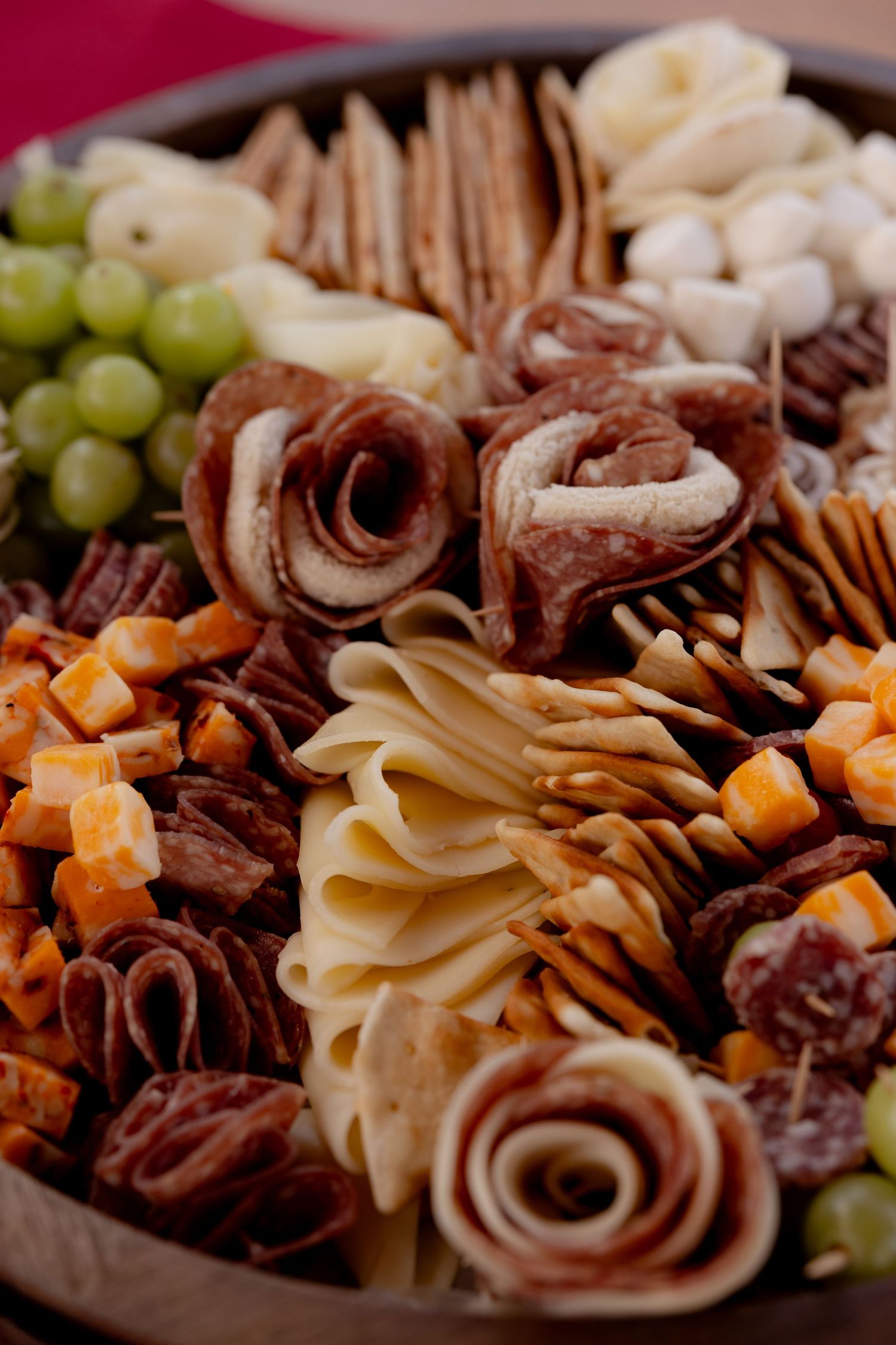 Close-up of charcuterie board with rolled meats, cheese, grapes, and nuts arranged artfully on dark platter.