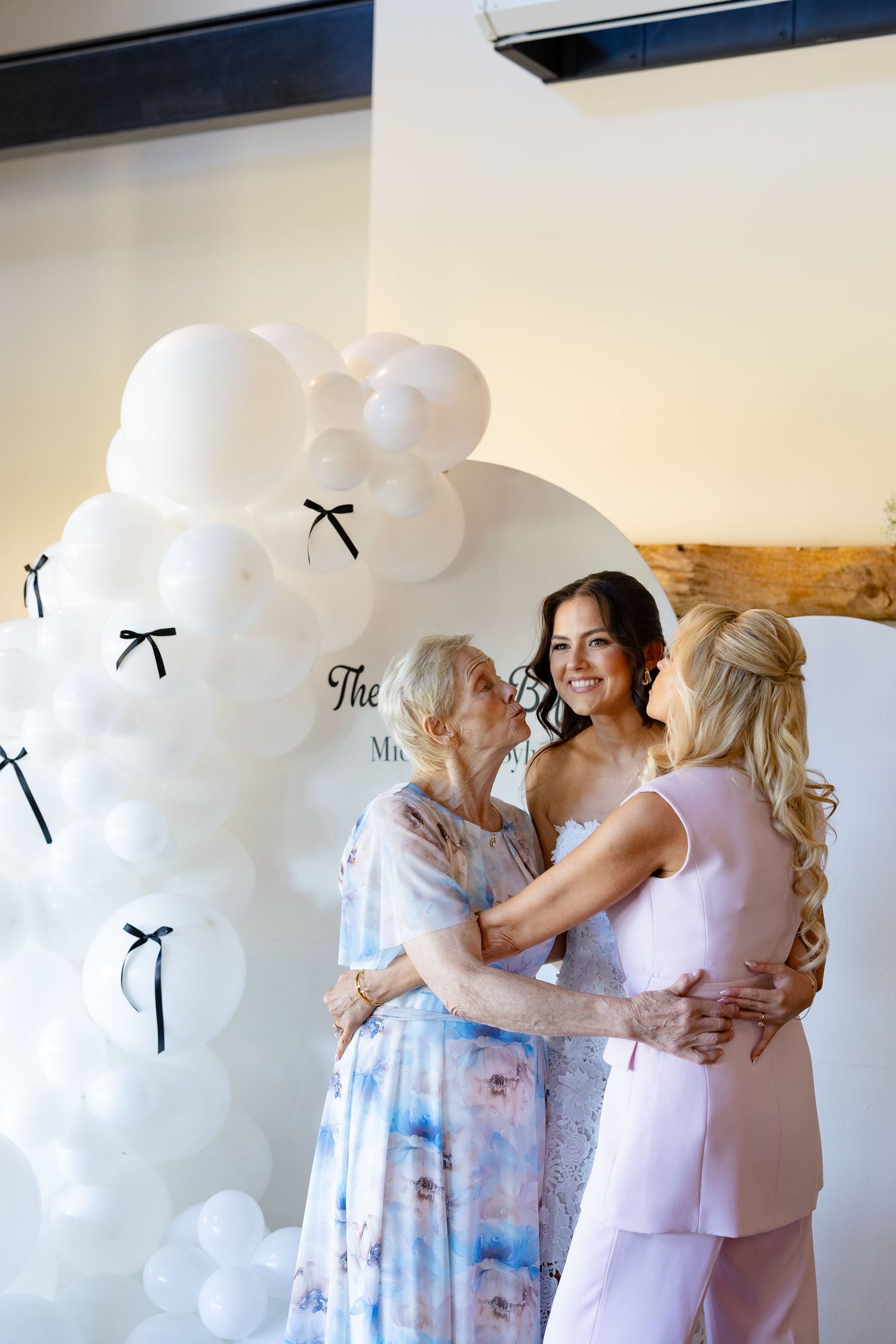 A group shares emotional embraces in front of a white balloon arch at a wedding celebration.