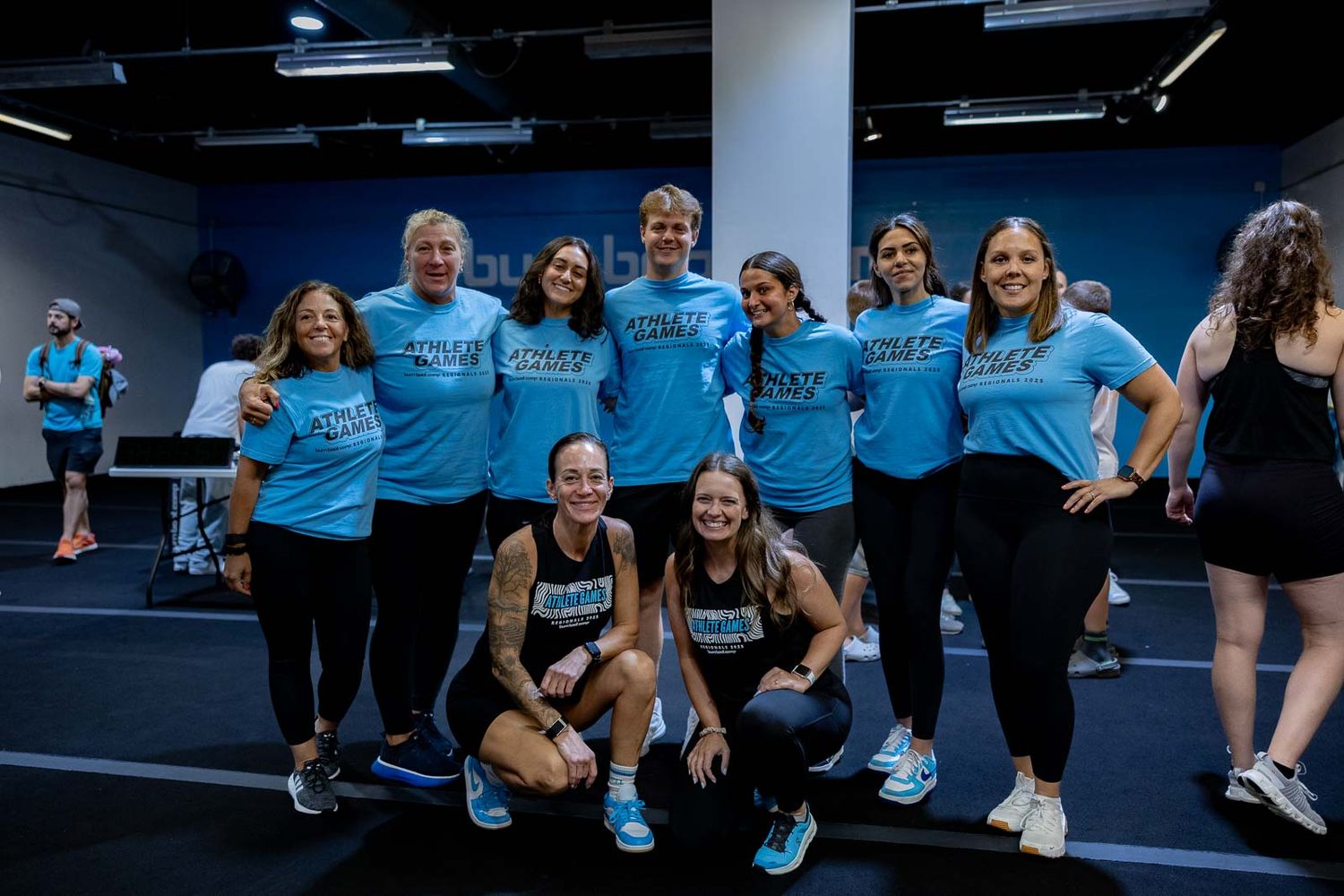 A group wearing matching light blue t-shirts poses together in an indoor exercise or dance studio space.