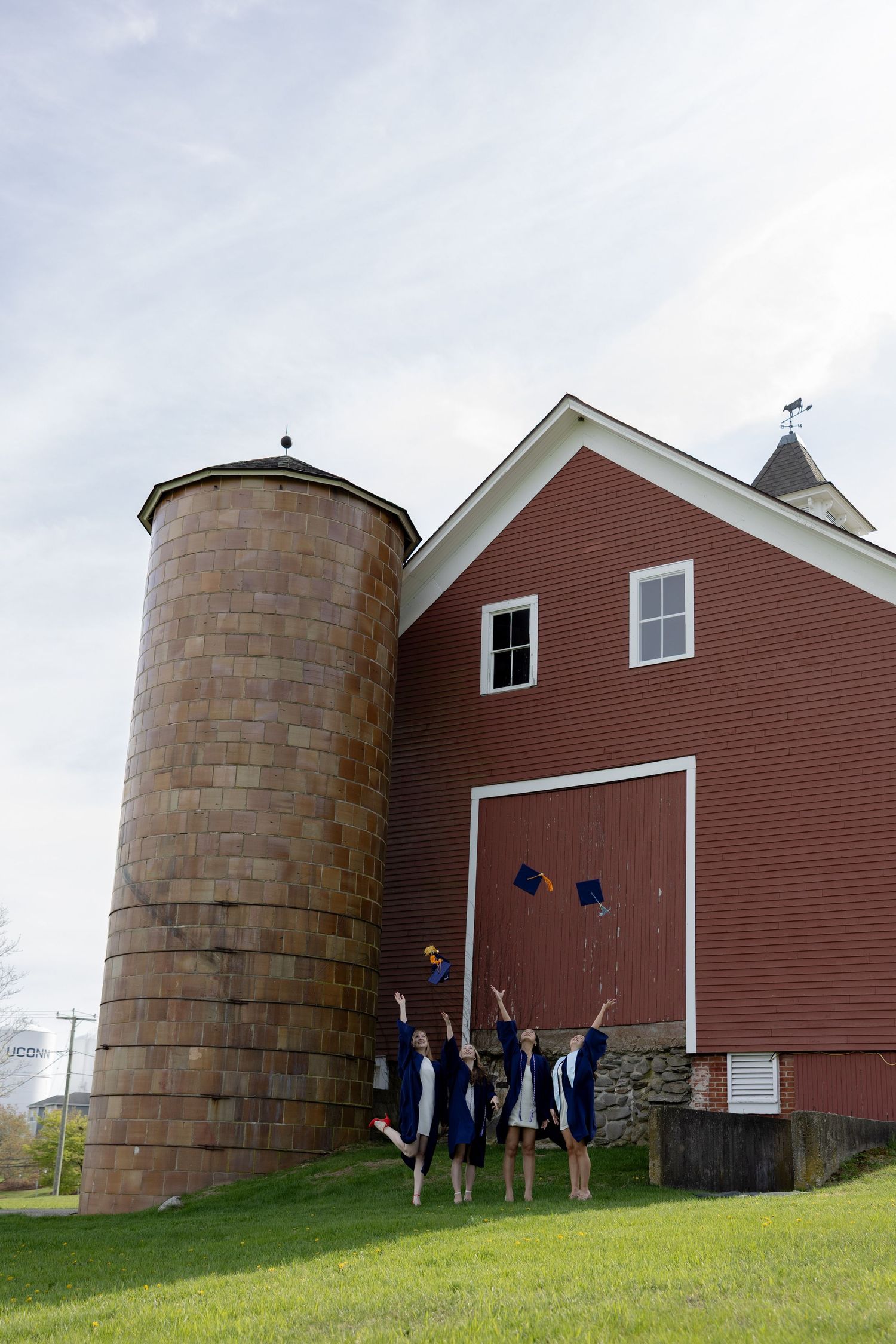 Graduates celebrate by throwing caps in the air in front of a classic red barn and stone silo on a grassy hillside.