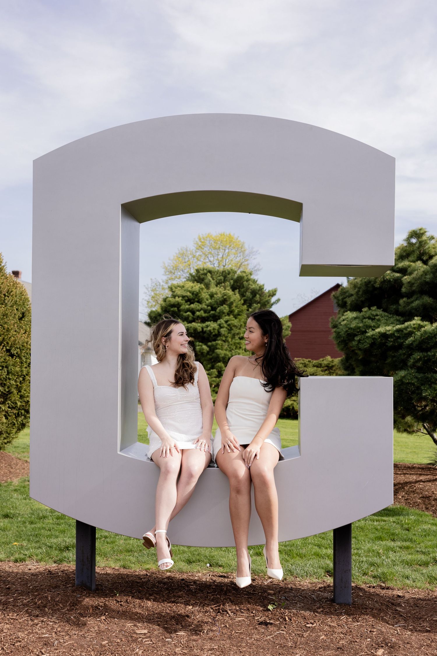 Two students in white dresses sit together in a giant letter G sculpture outdoors on a college campus.
